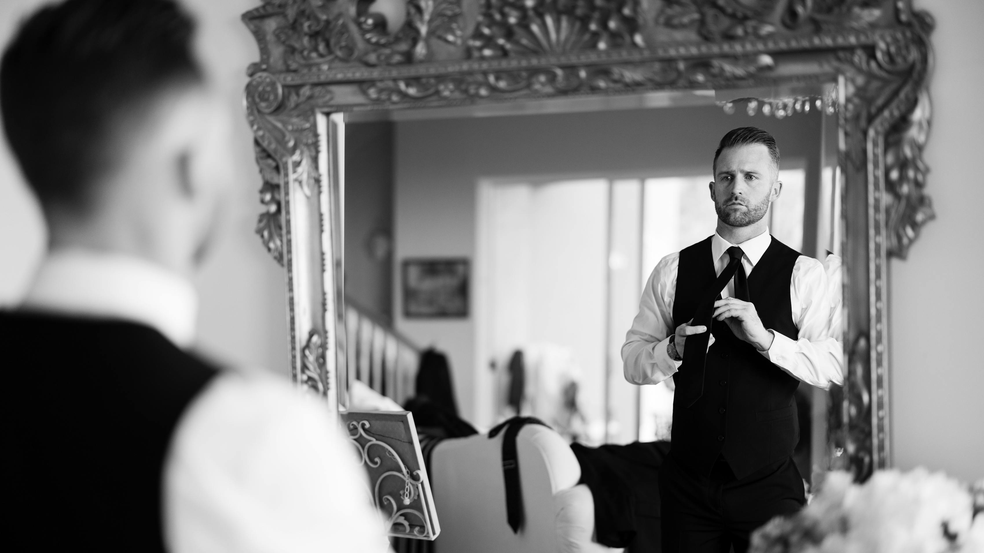 Man getting dressed to be a wedding guest tying neck tie in the mirror to match his black suit vest and suit pants.