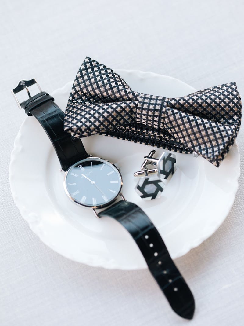 Men's cocktail attire suit accessories with black leather watch, black and white cufflinks, and patterned bow tie.
