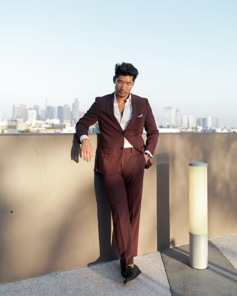 Men's burgundy suit with unbuttoned white dress shirt, pink pocket square, and black loafers in front of city skyline.