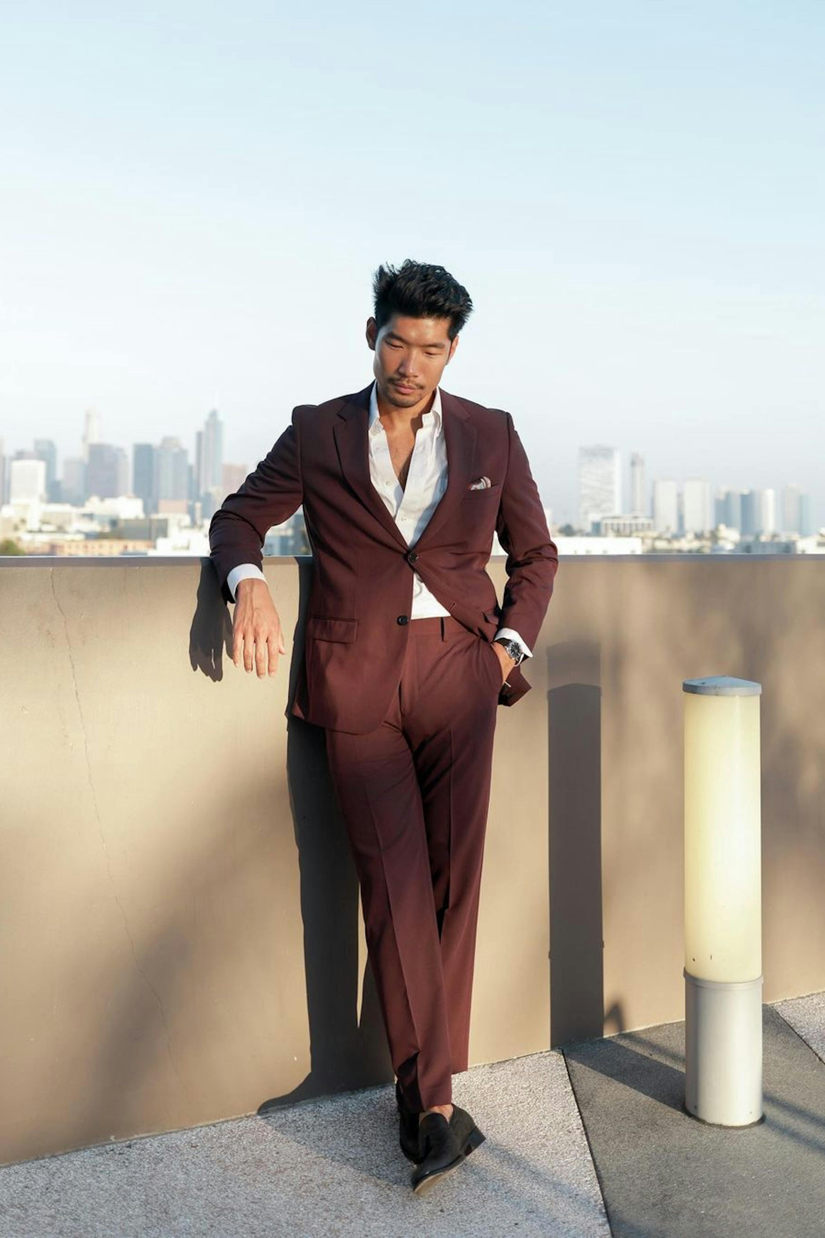Men's burgundy suit with unbuttoned white dress shirt, pink pocket square, and black loafers in front of city skyline.