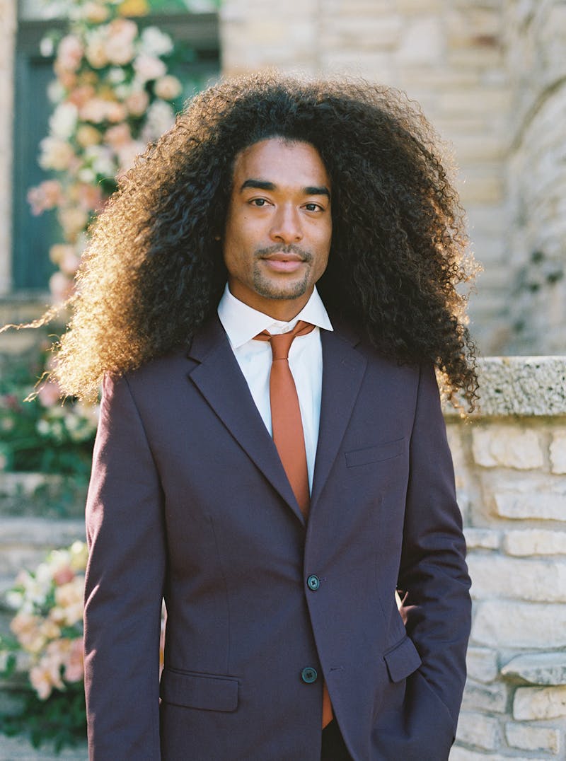 Long haired man wearing deep maroon burgundy suit with bold rust tie at a wedding.