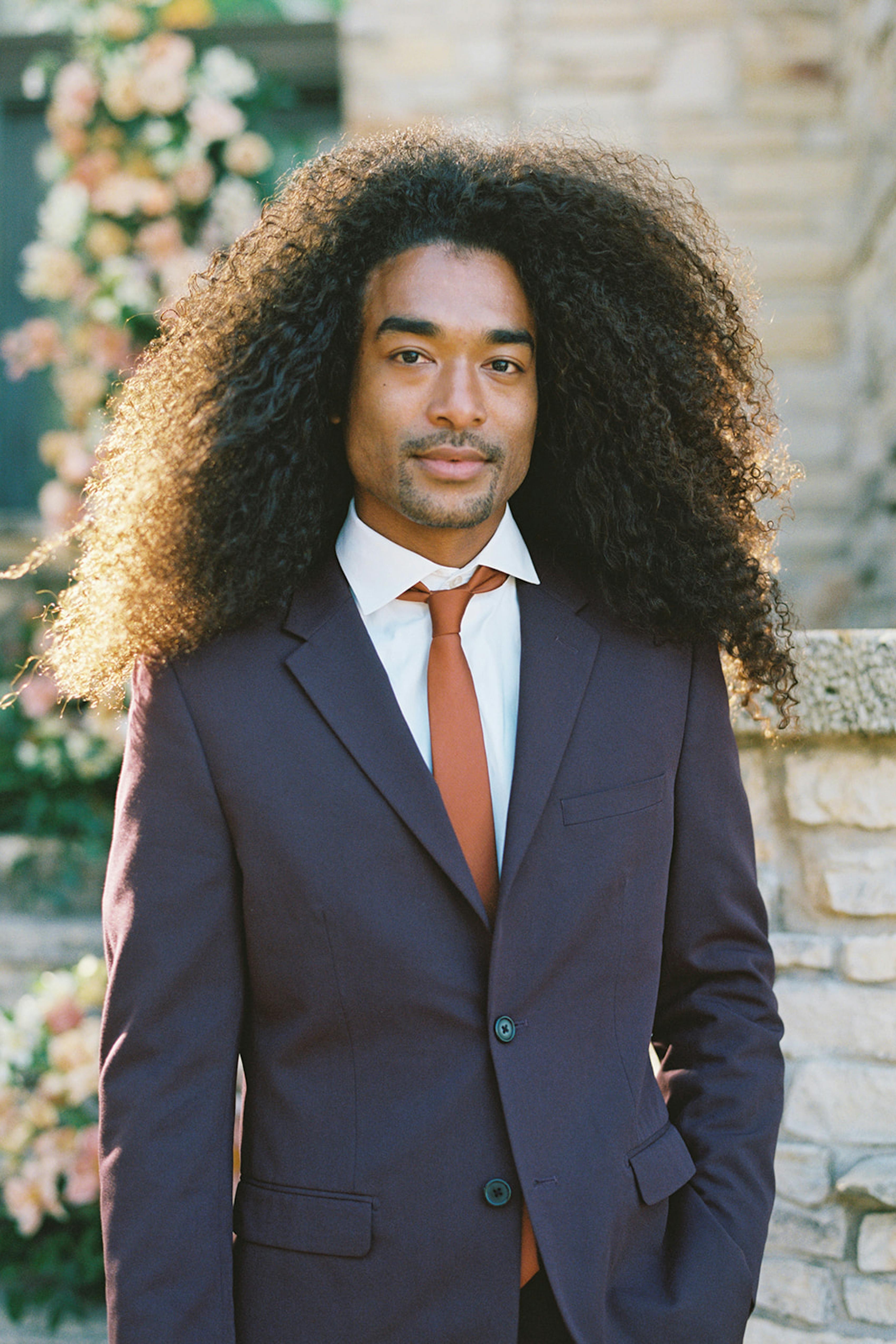 Long haired man wearing deep maroon burgundy suit with bold rust tie at a wedding.