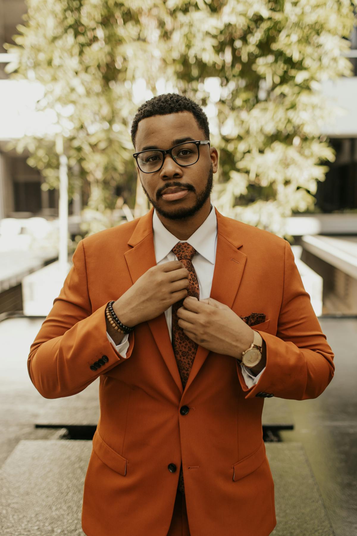 Rust orange suit outfit for cocktail dress code wedding with tie, watch, and glasses.