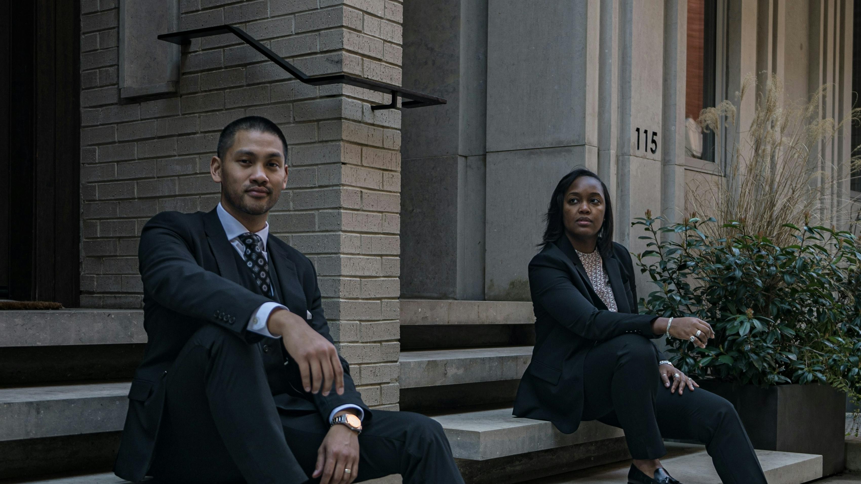 A man and a women in dark suits for professional outfits sitting on office steps showing off dress shoes.