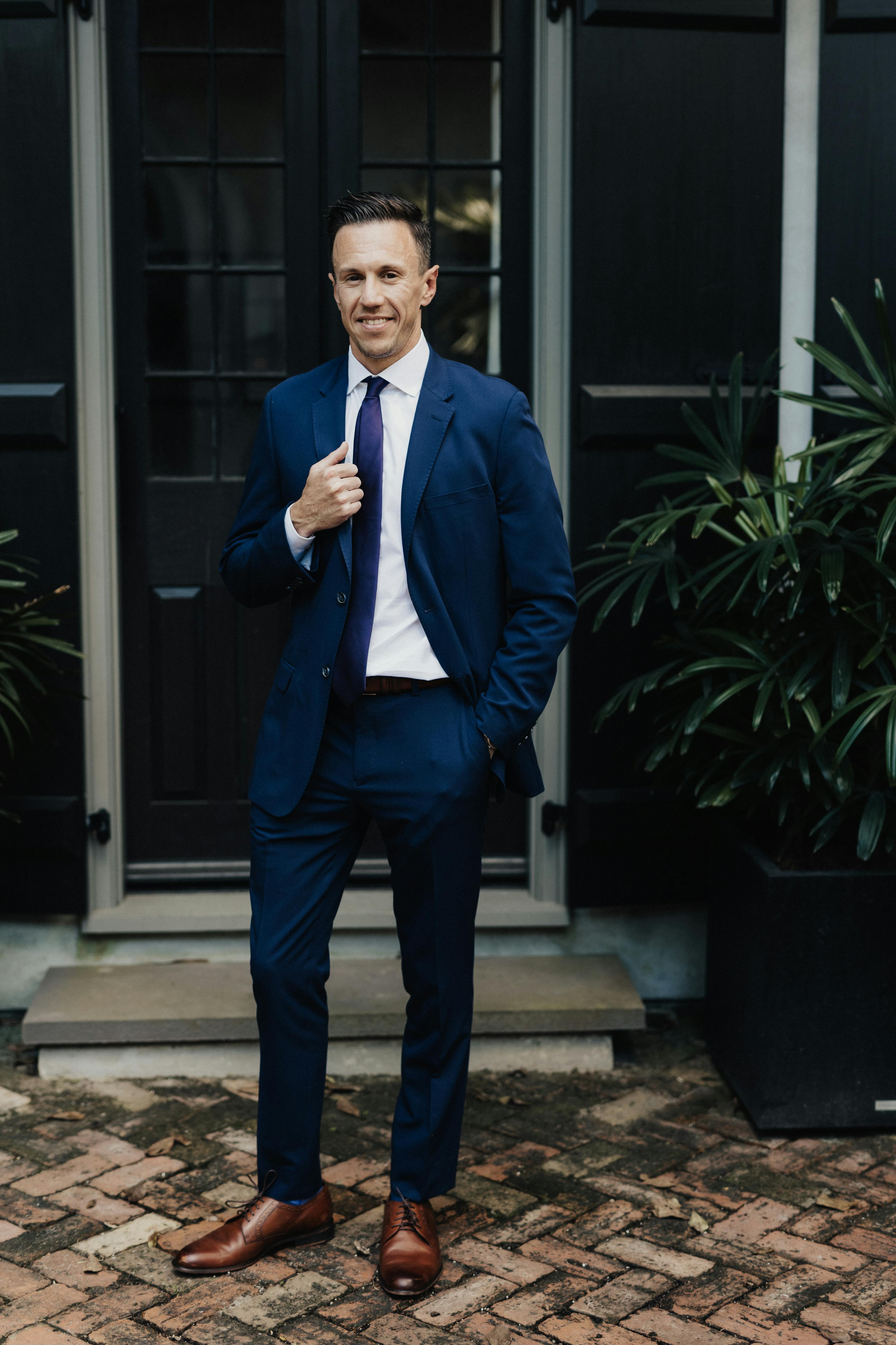Man wearing formal dress code wedding guest outfit with navy blue suit and tie and brown shoes.