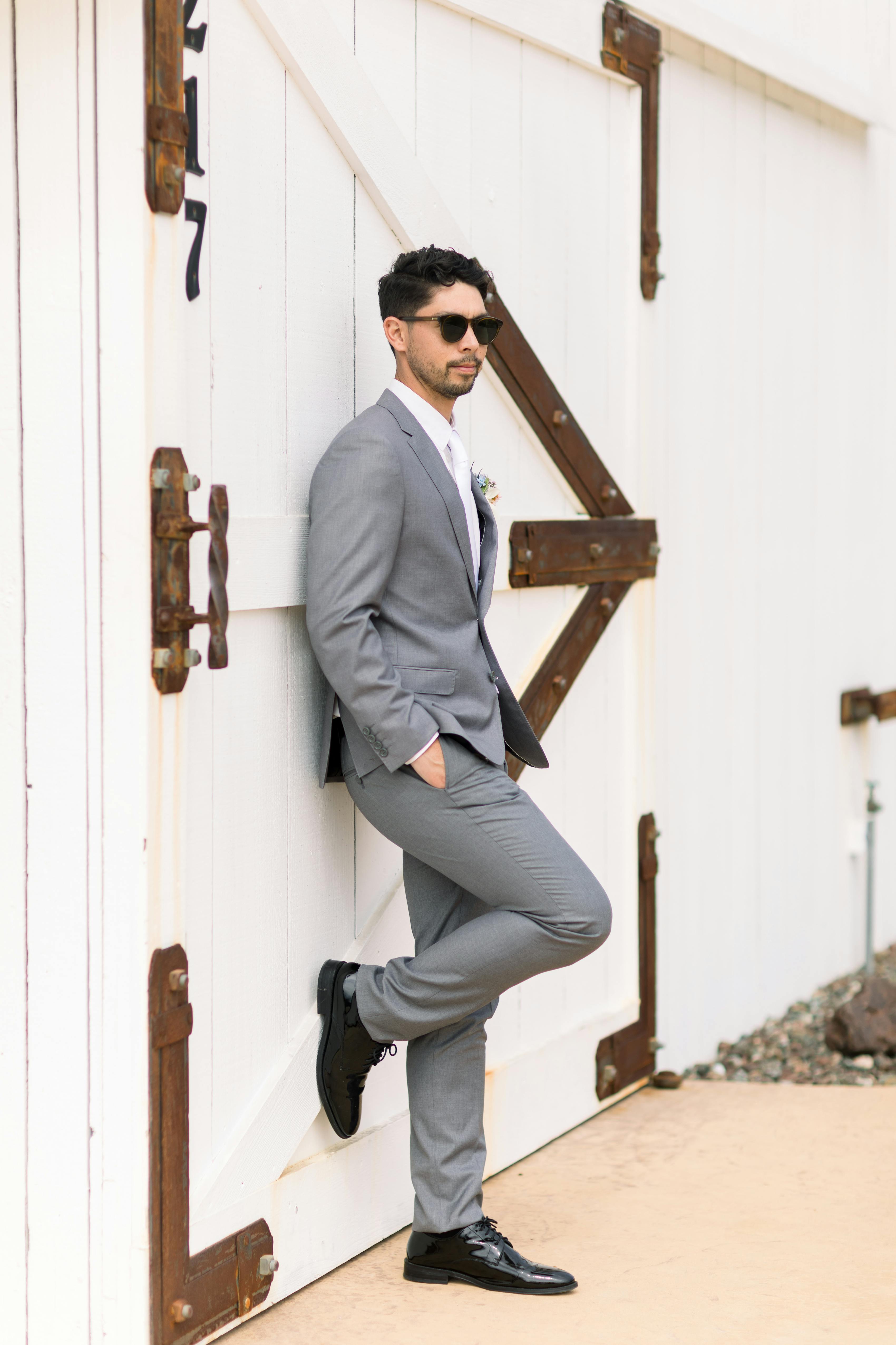 Man posing outside in sunglasses and light grey suit with black shoes.