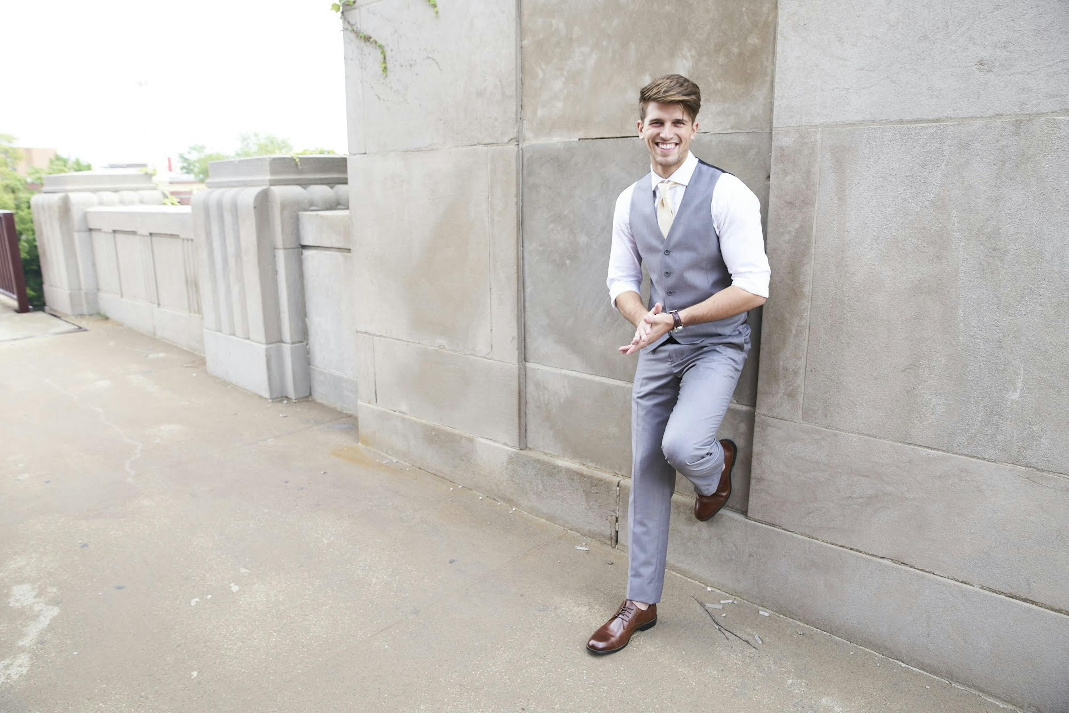 Man wearing light gray suit vest and grey suit pants with dark brown shoes posing outside at wedding