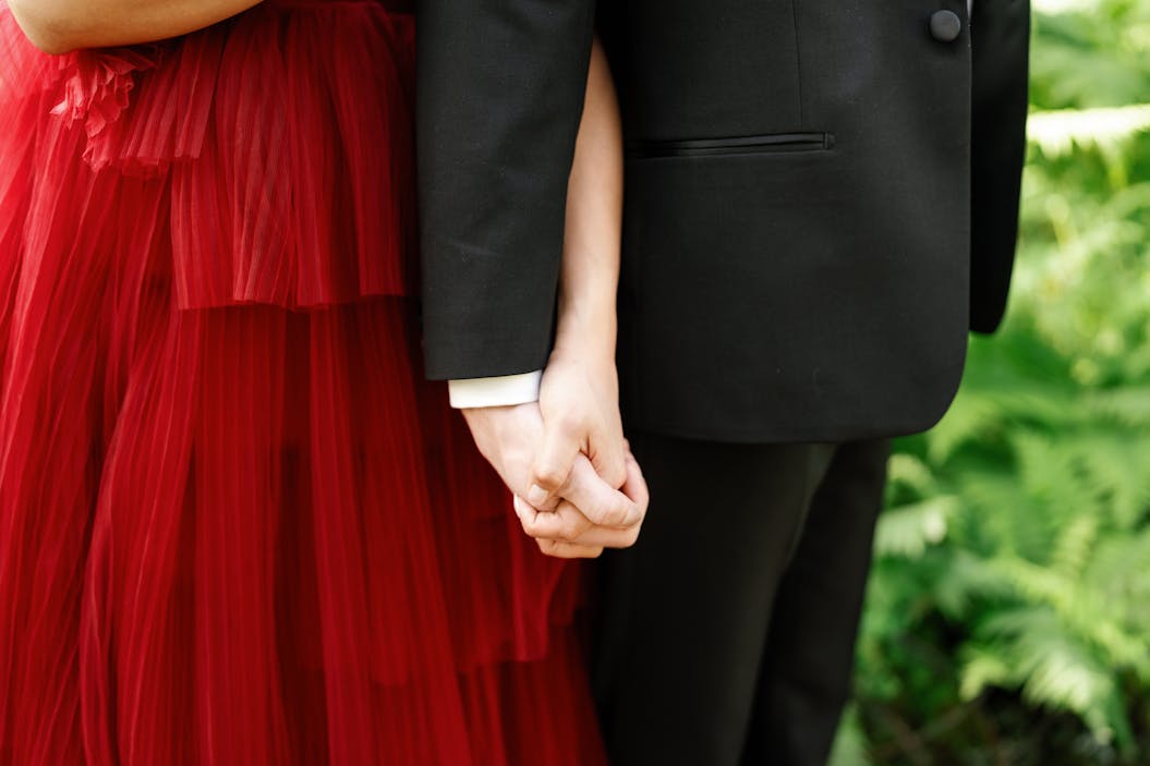 Wedding guests holding hands wearing women's red gown and men's black tuxedos for formal attire.