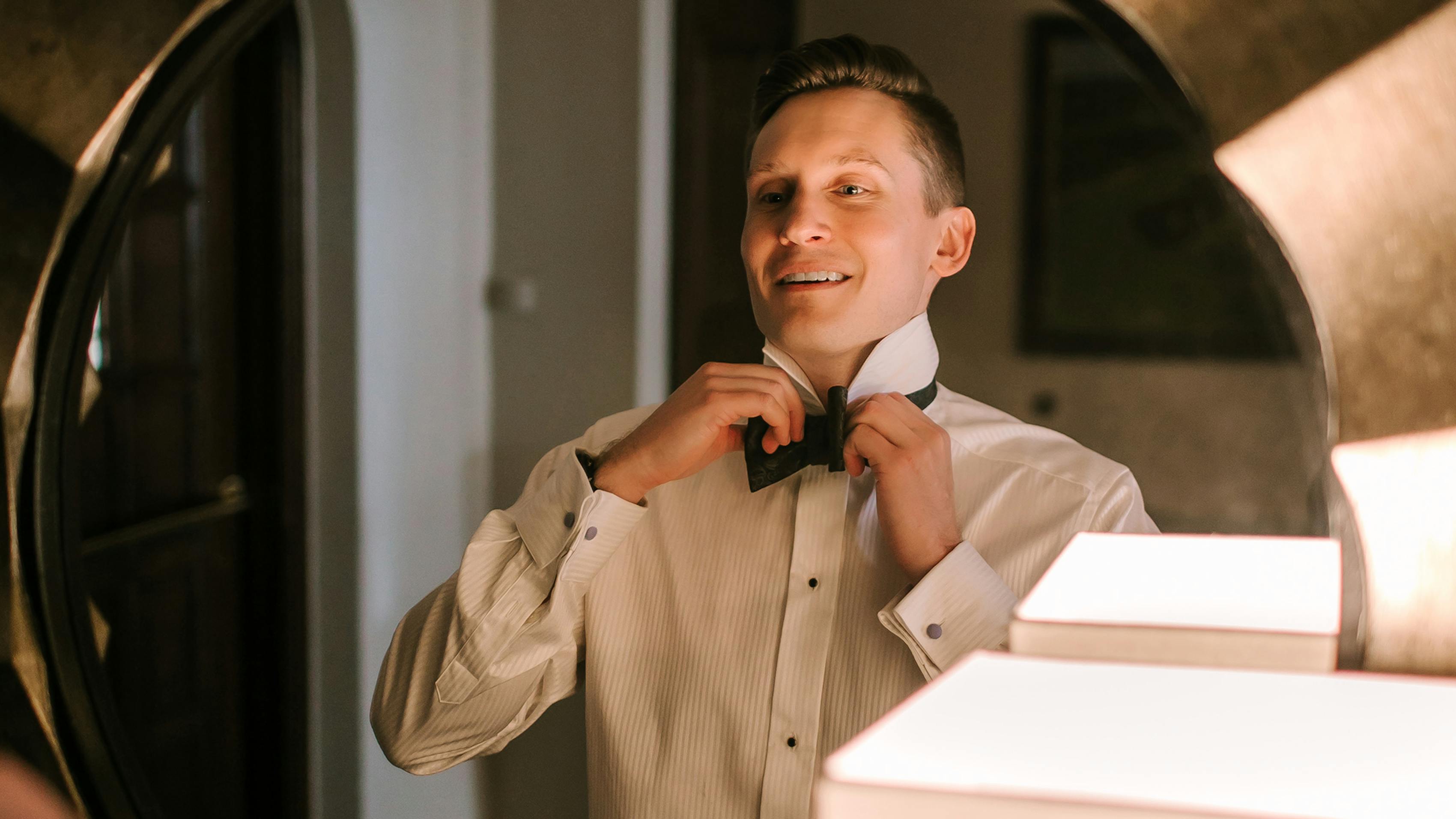 Groom gets ready for his wedding day by tying his bow tie in front of a mirror.