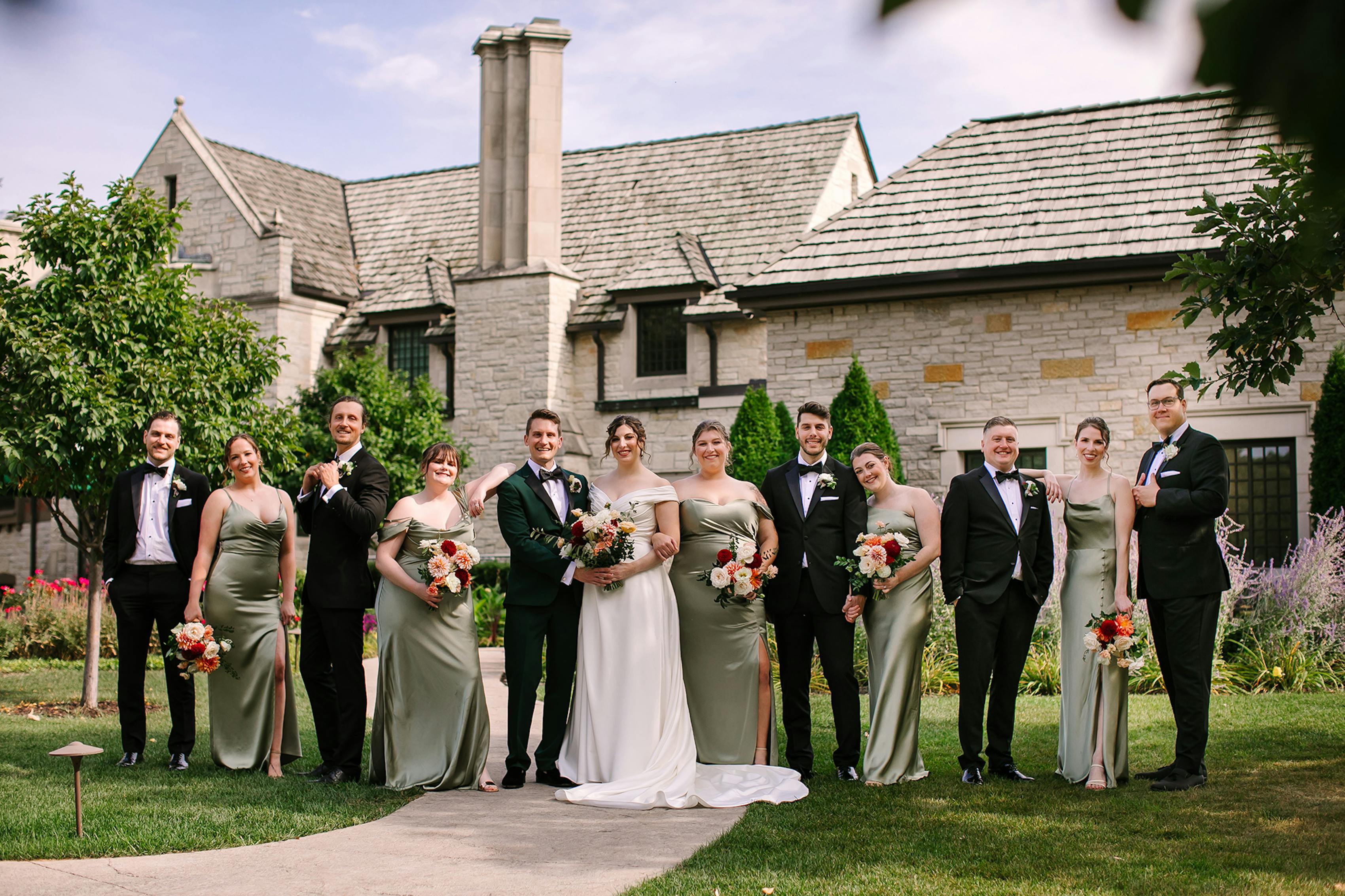 Wedding party dressed in black tuxedos and sage green bridesmaids dresses with the groom standing out in a green tuxedo and the bride wearing white.