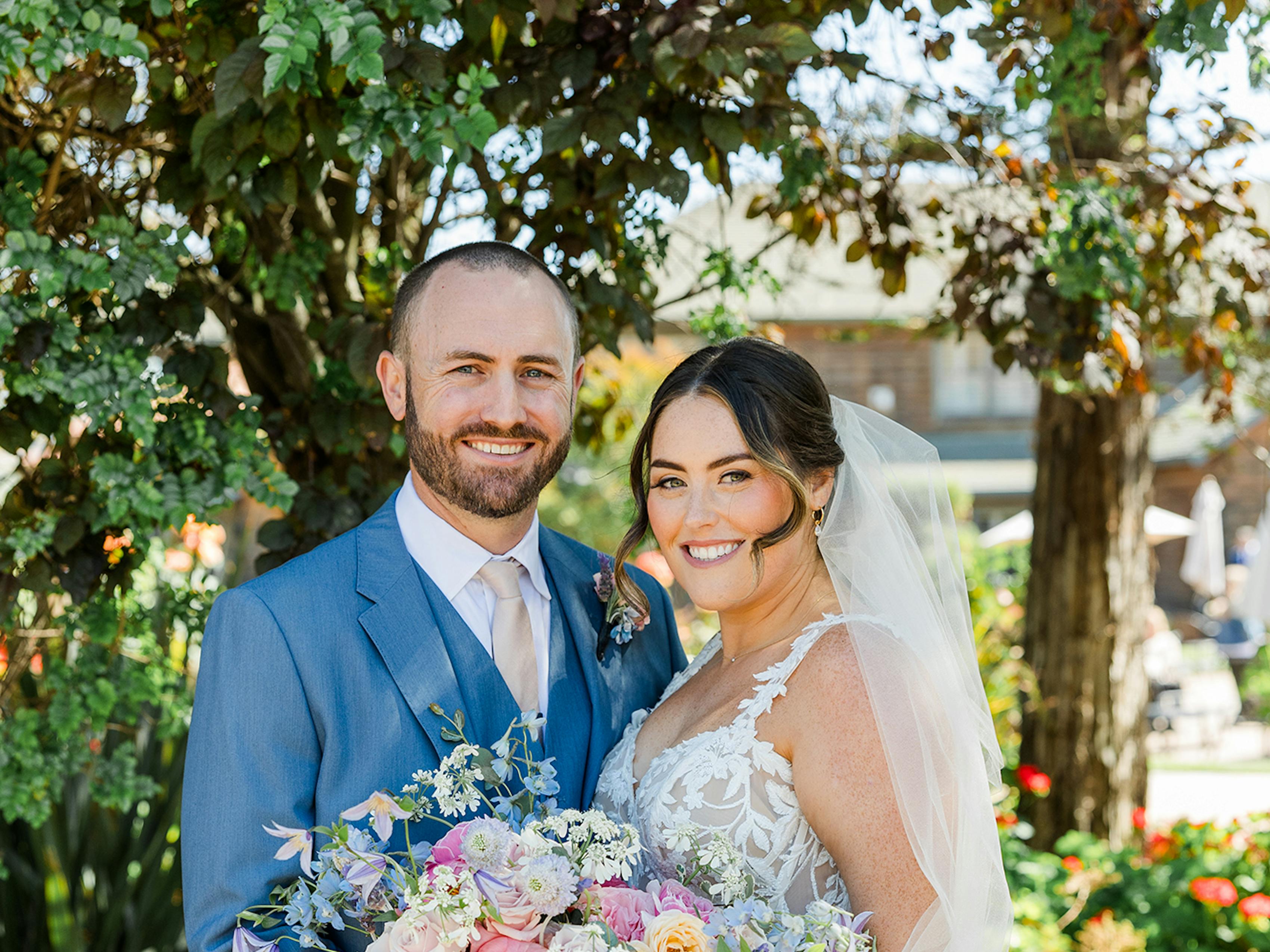 Bride and groom pose in their spring wedding attire with a large bridal bouquet of pink and yellow roses.