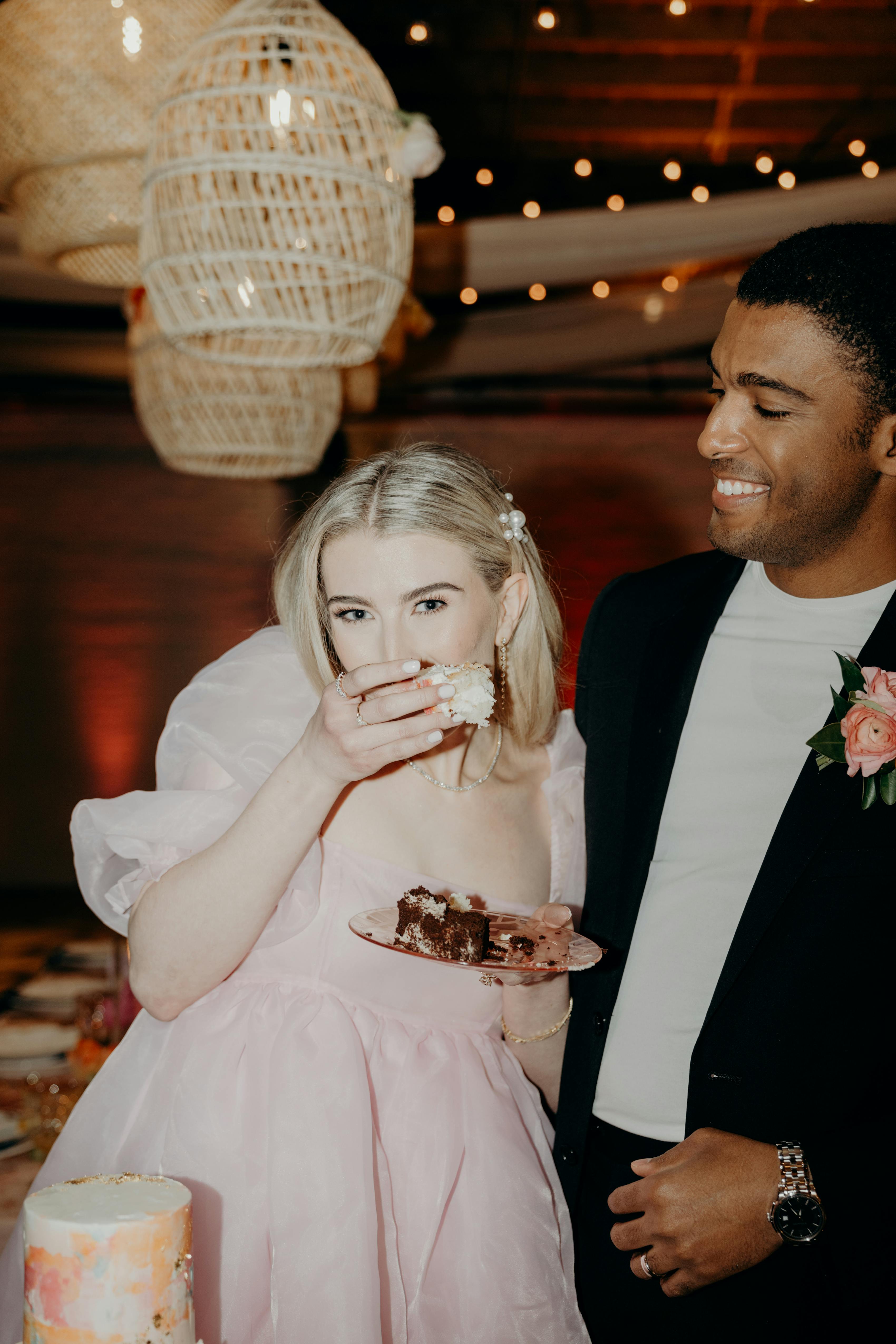 Bride eating cake in trendy pink short wedding dress beside groom laughing in casual black wedding suit with t shirt