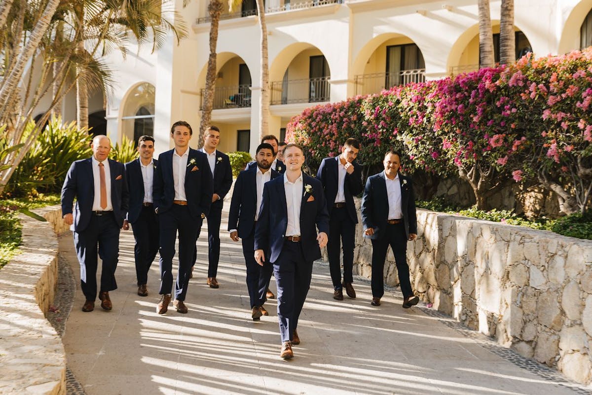 Groomsmen wearing blue suits for men with white dress shirts and no tie walking to beach wedding