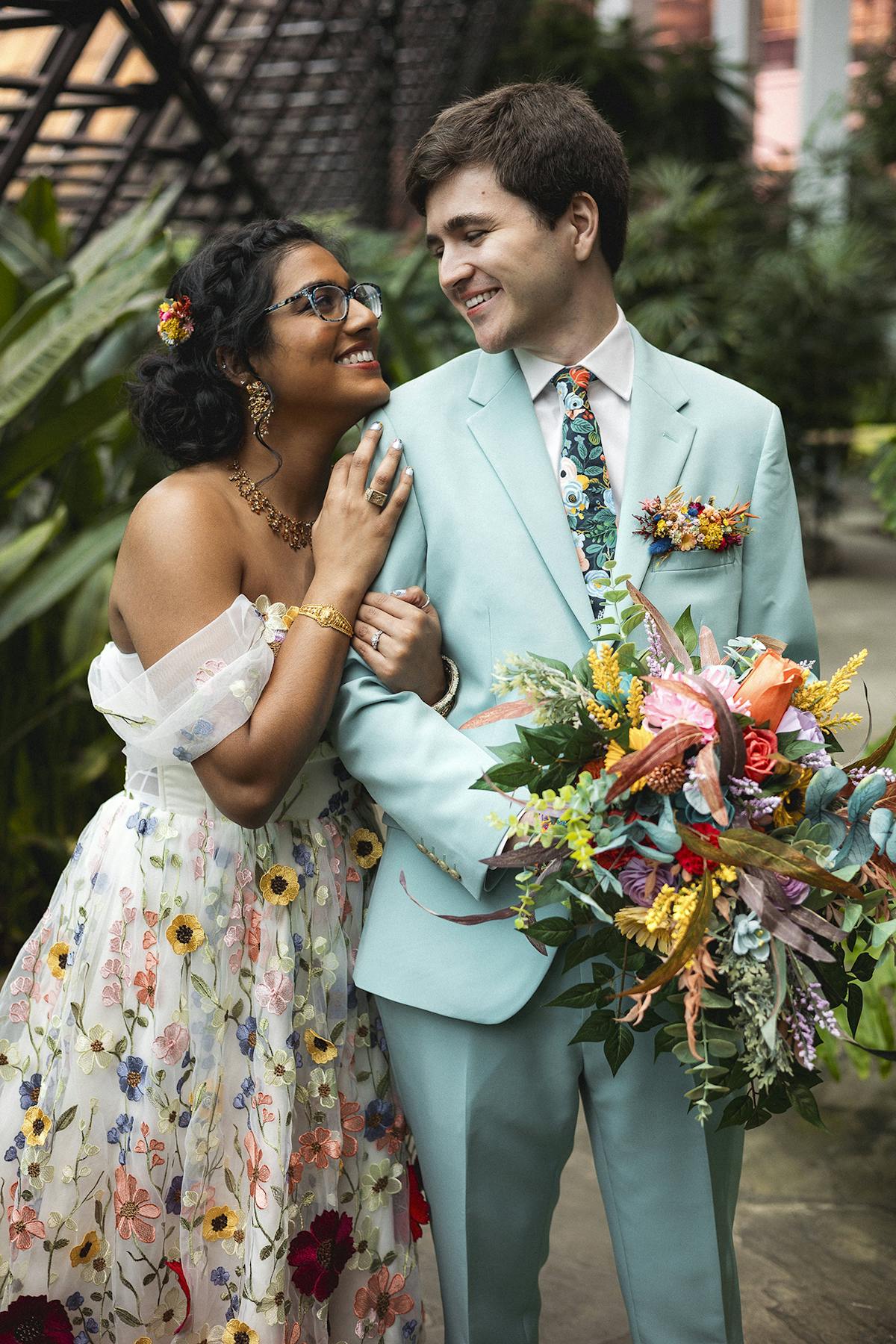 Bride wears floral dress to colorful wedding while groom looks lovingly in a seaglass suit for men