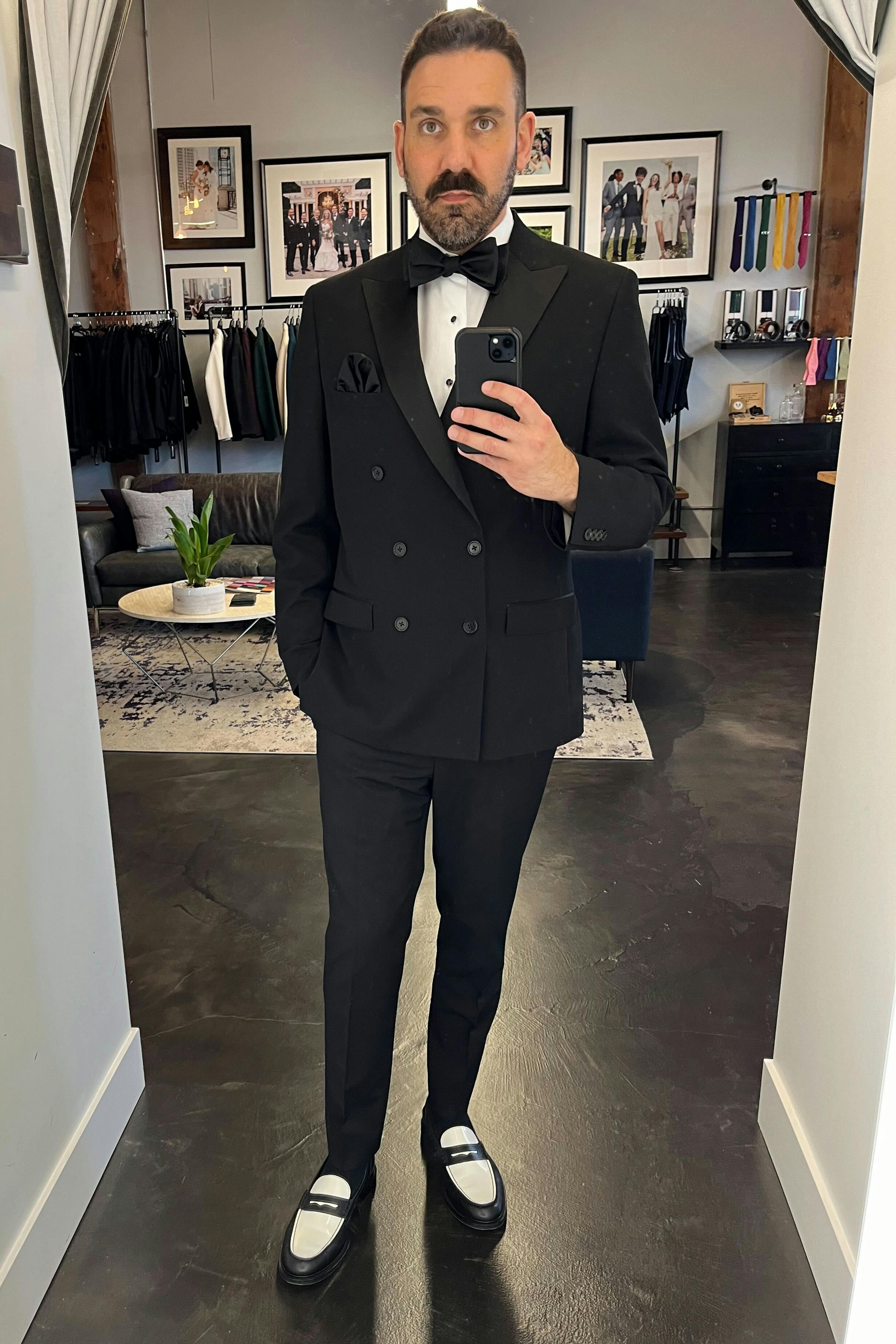 Man taking mirror selfie wearing double breasted peak lapel black tuxedo and black tie accessories
