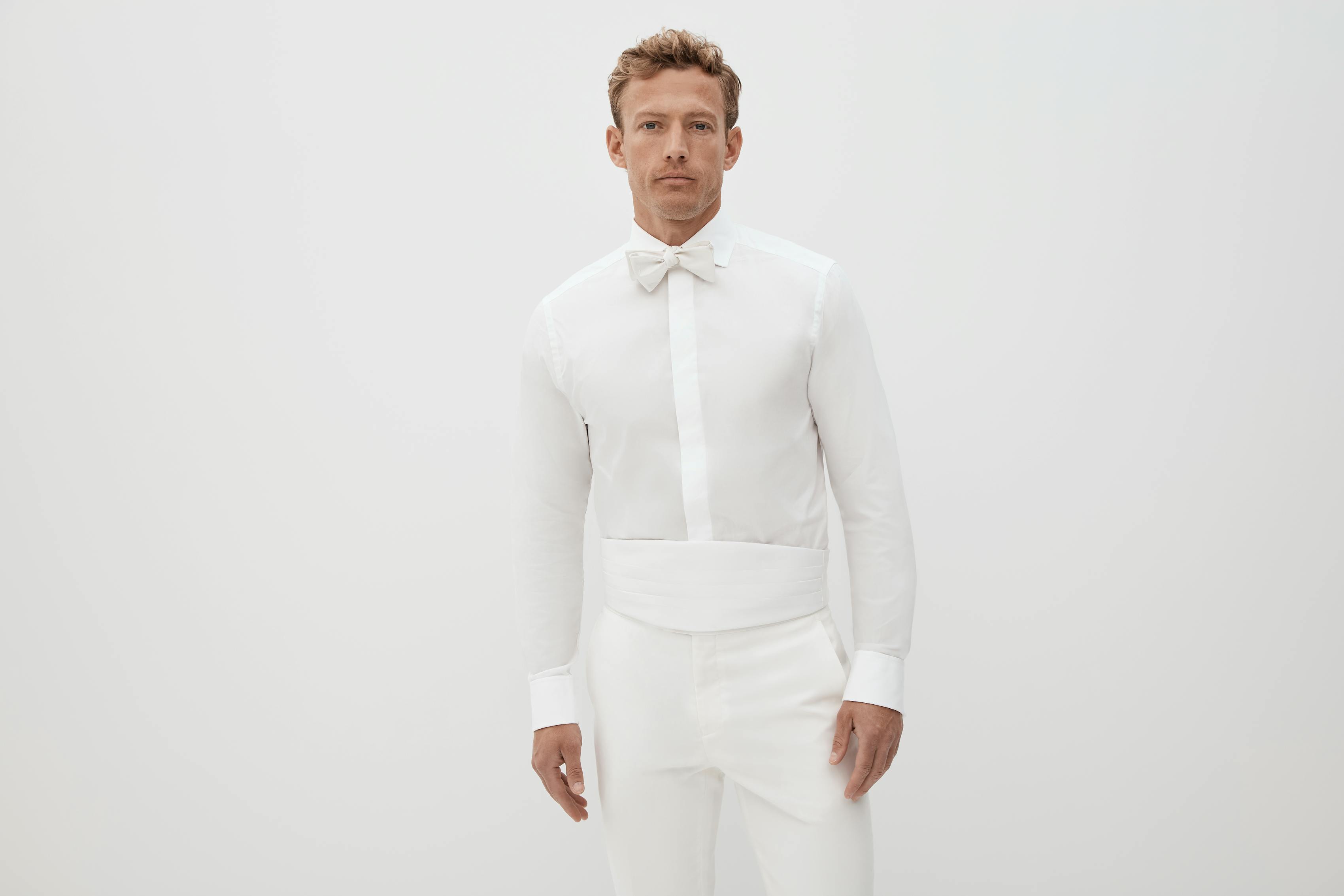 Man getting ready for a white tie attire event with a French cuff shirt, white bow tie, and white tuxedo pants