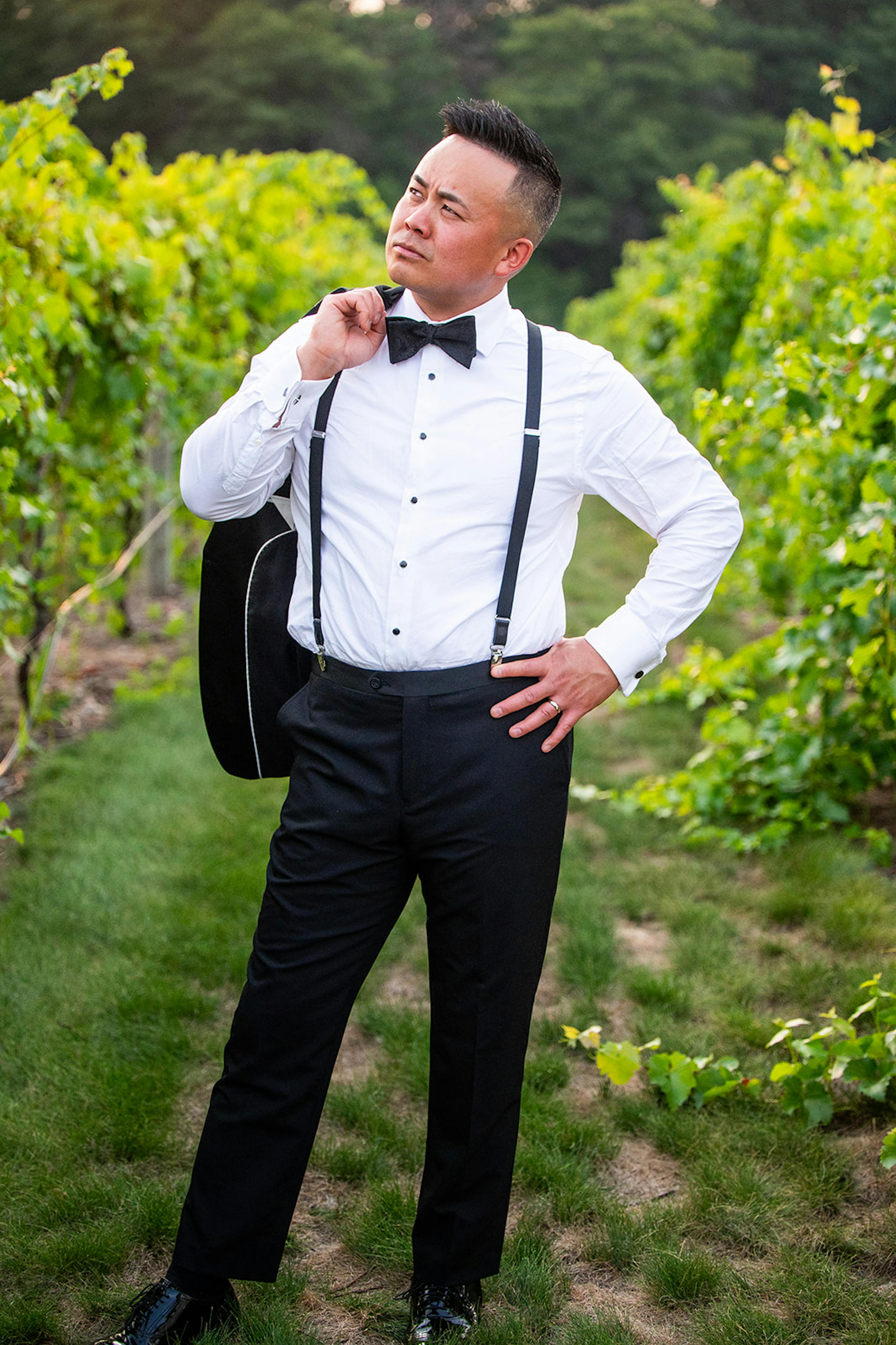 Black tie summer wedding guest wearing men's black tuxedo and suspenders at vineyard venue