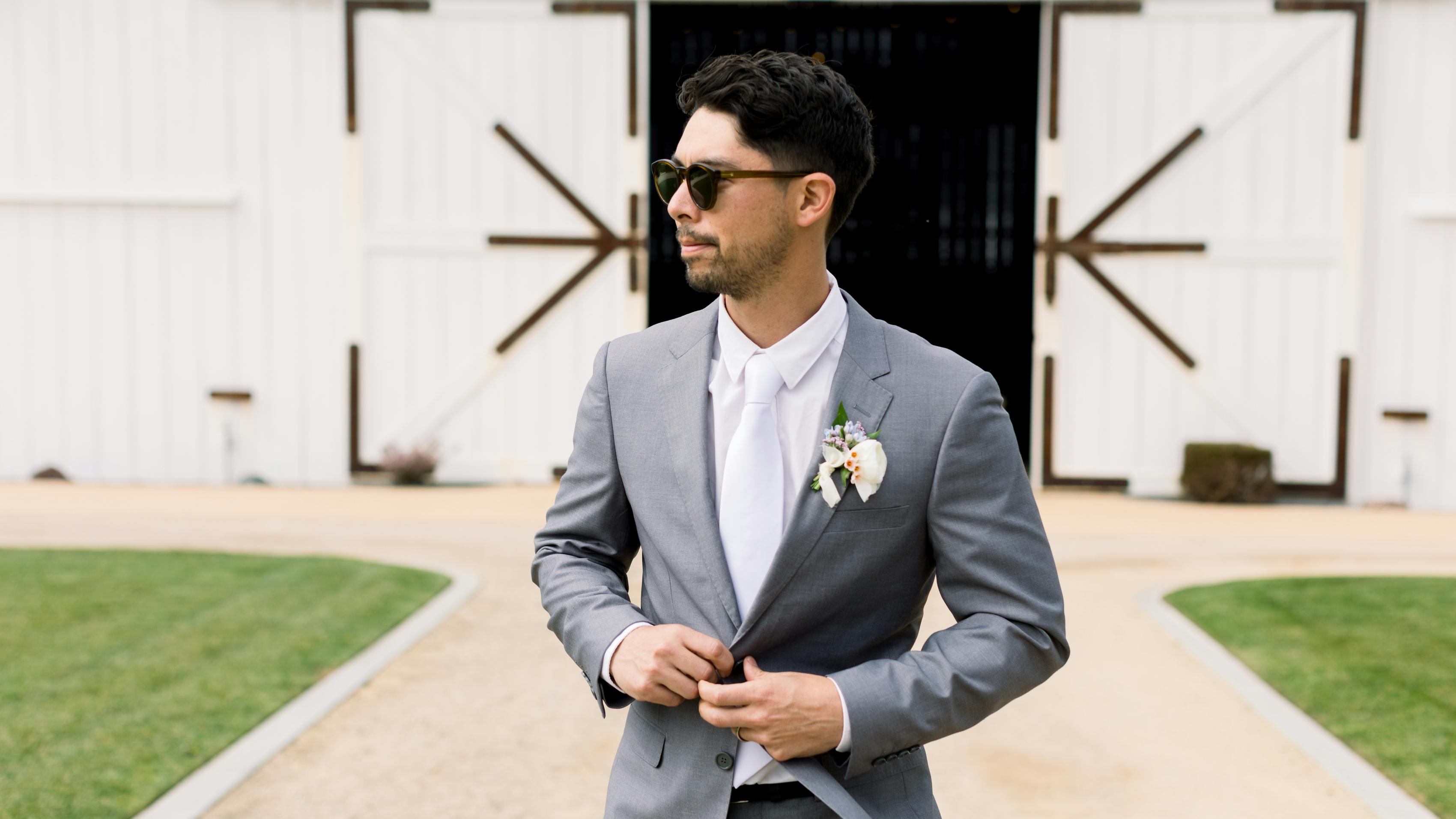 summer wedding guest wearing men's light grey suit, white suit accessories, and sunglasses at barn venue