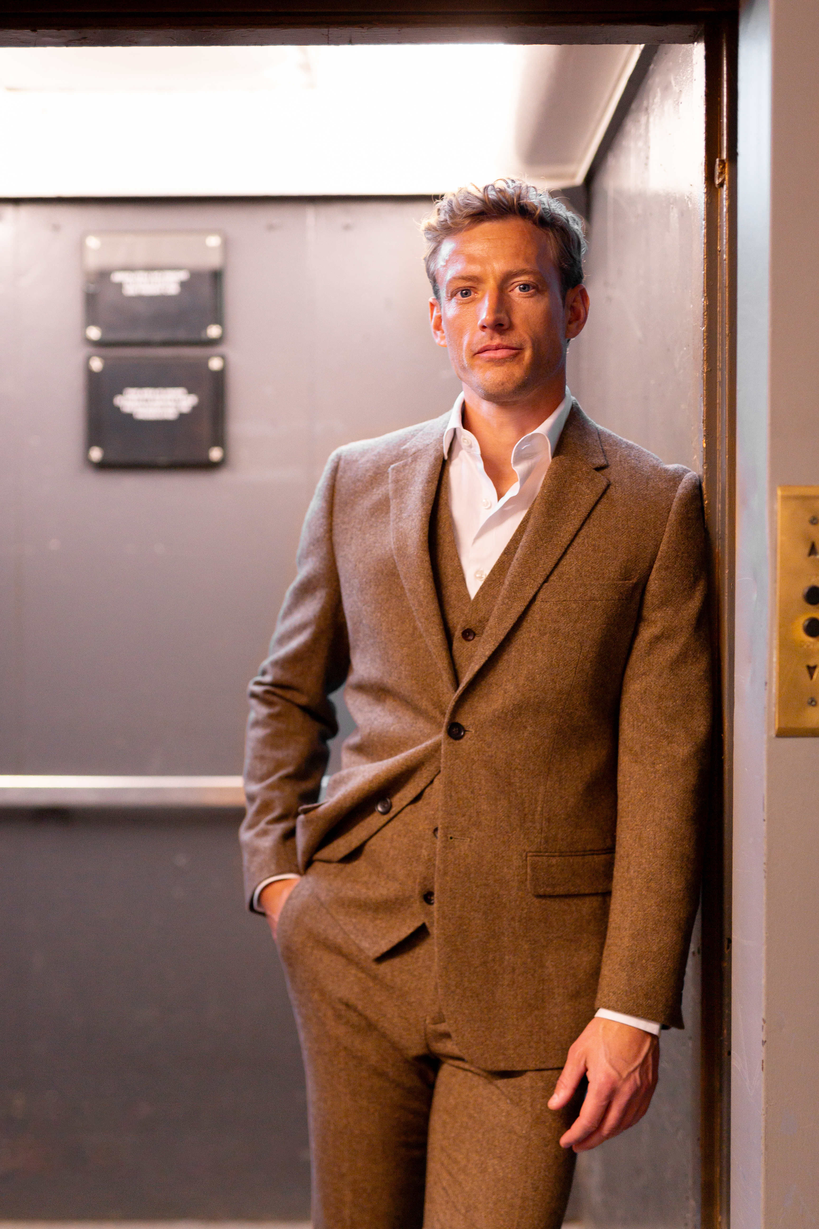 Man in elevator wearing simple, formal tweed suit with tweed vest and white dress shirt.