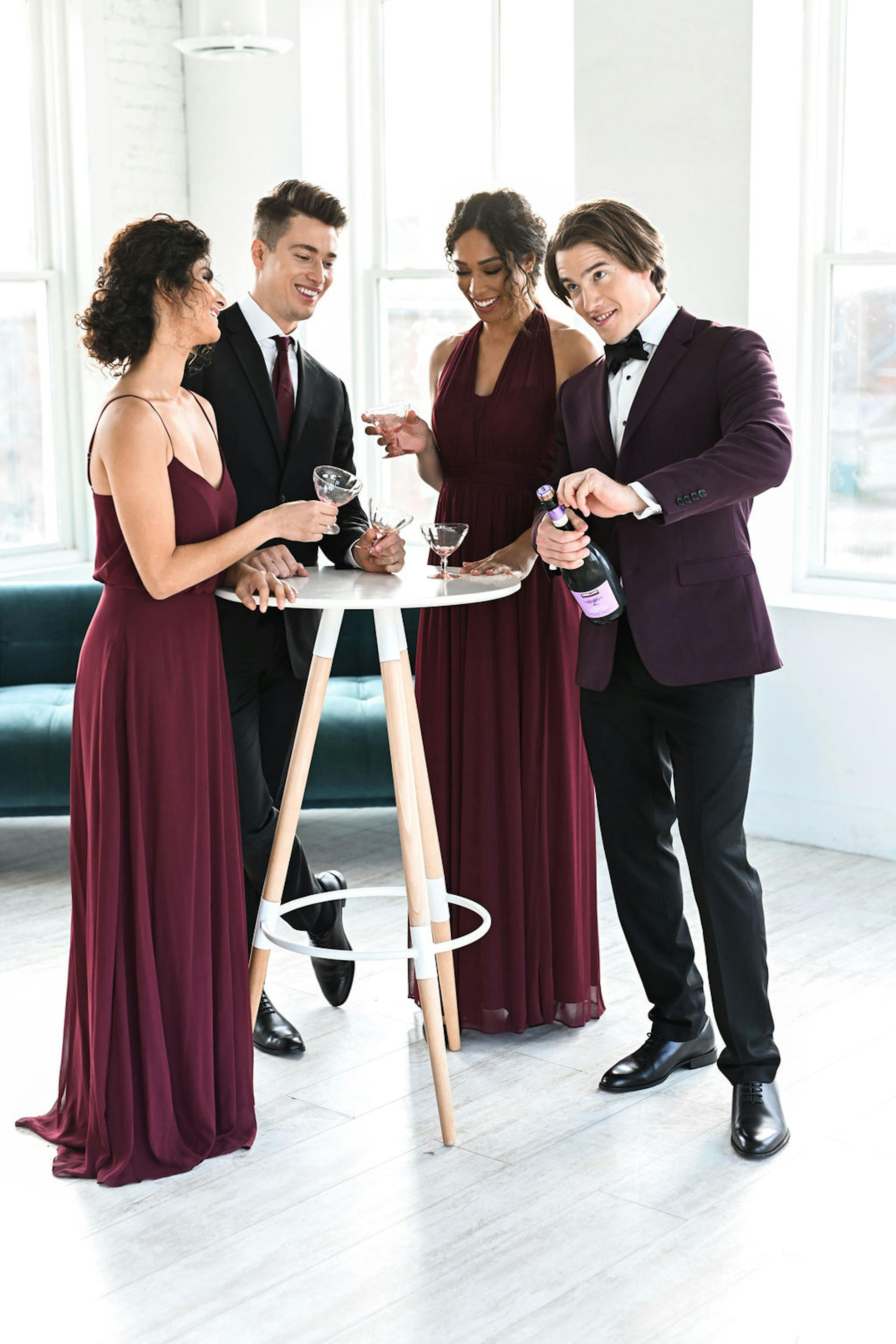A group of coordinated men and women wear burgundy tuxedos and floor length gowns to a festive holiday party