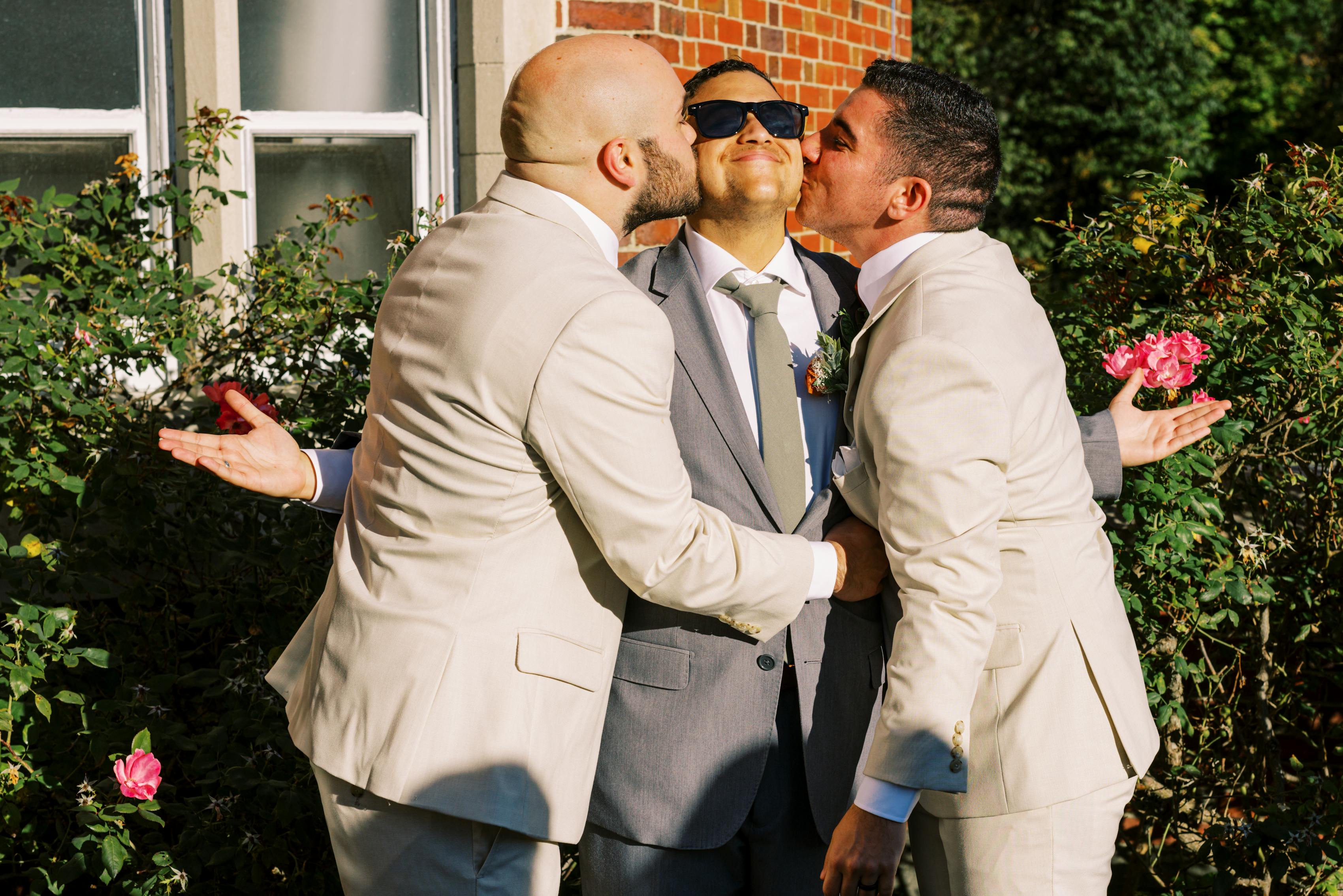 Two grooms in tan wedding suits, giving friend in gray groomsman suit a kiss on either cheeck