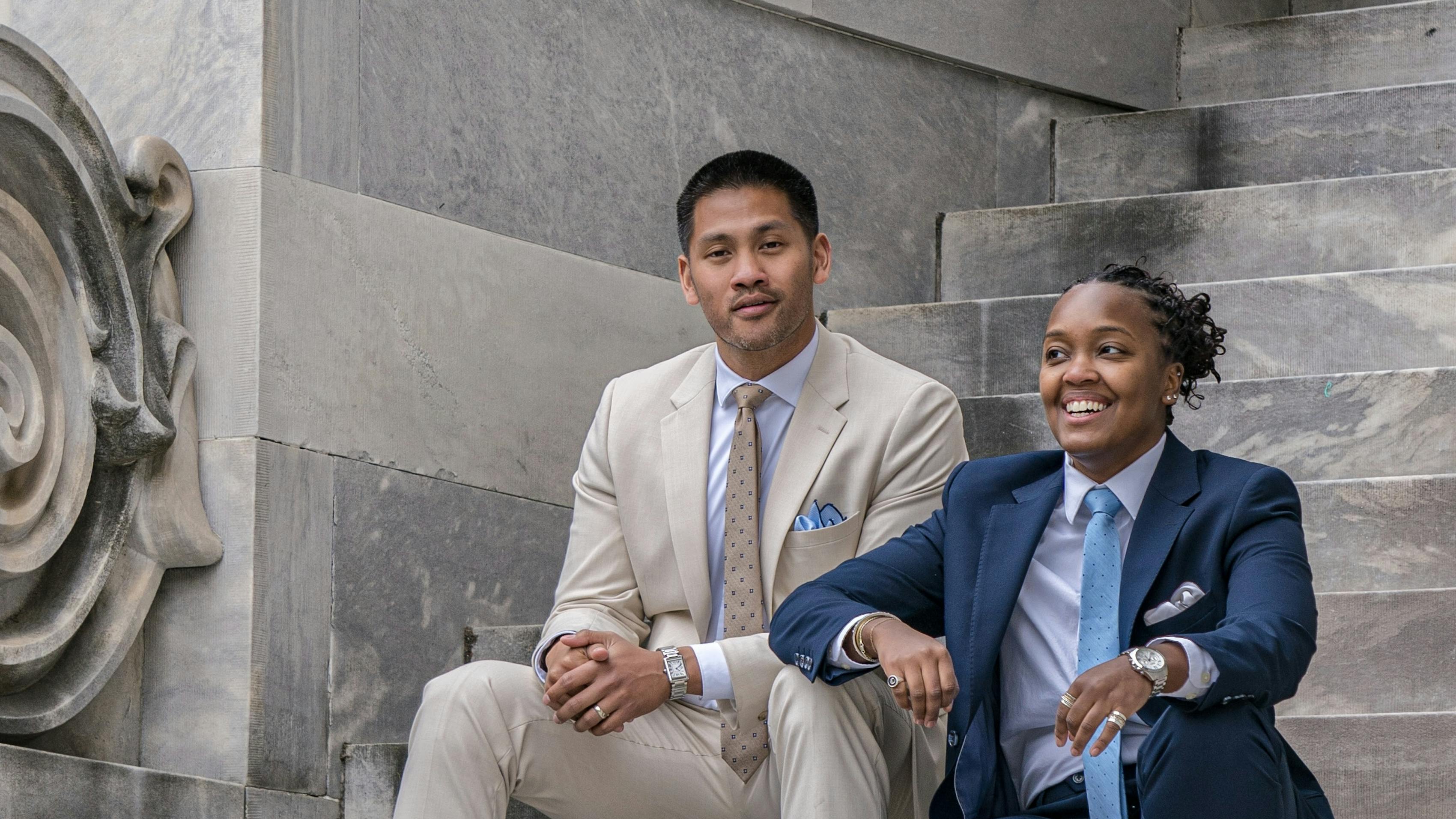 Man wearing light tan suit and woman wearing navy blue in front of professional building.