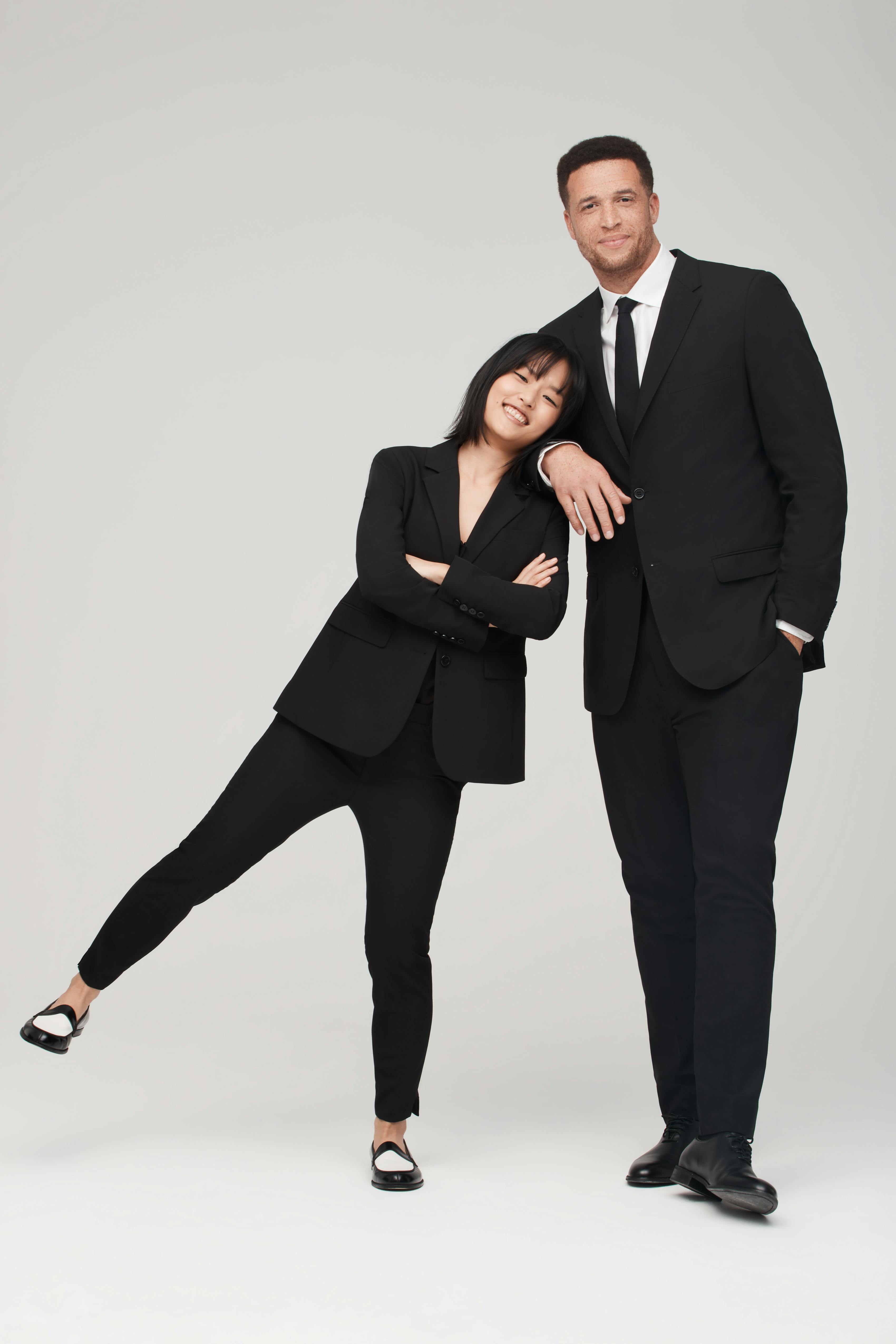 Short woman and tall man leaning against each other wearing matching black suits.