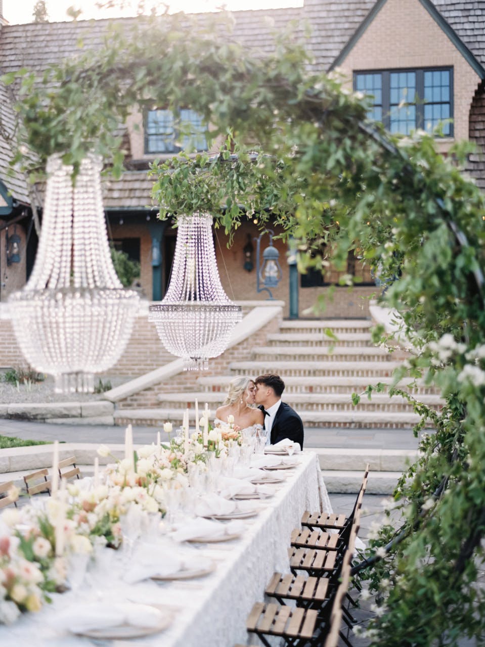 Bride and groom kiss at the end of a banquet table in that garden at modern European themed outdoor wedding with neutral color palette.