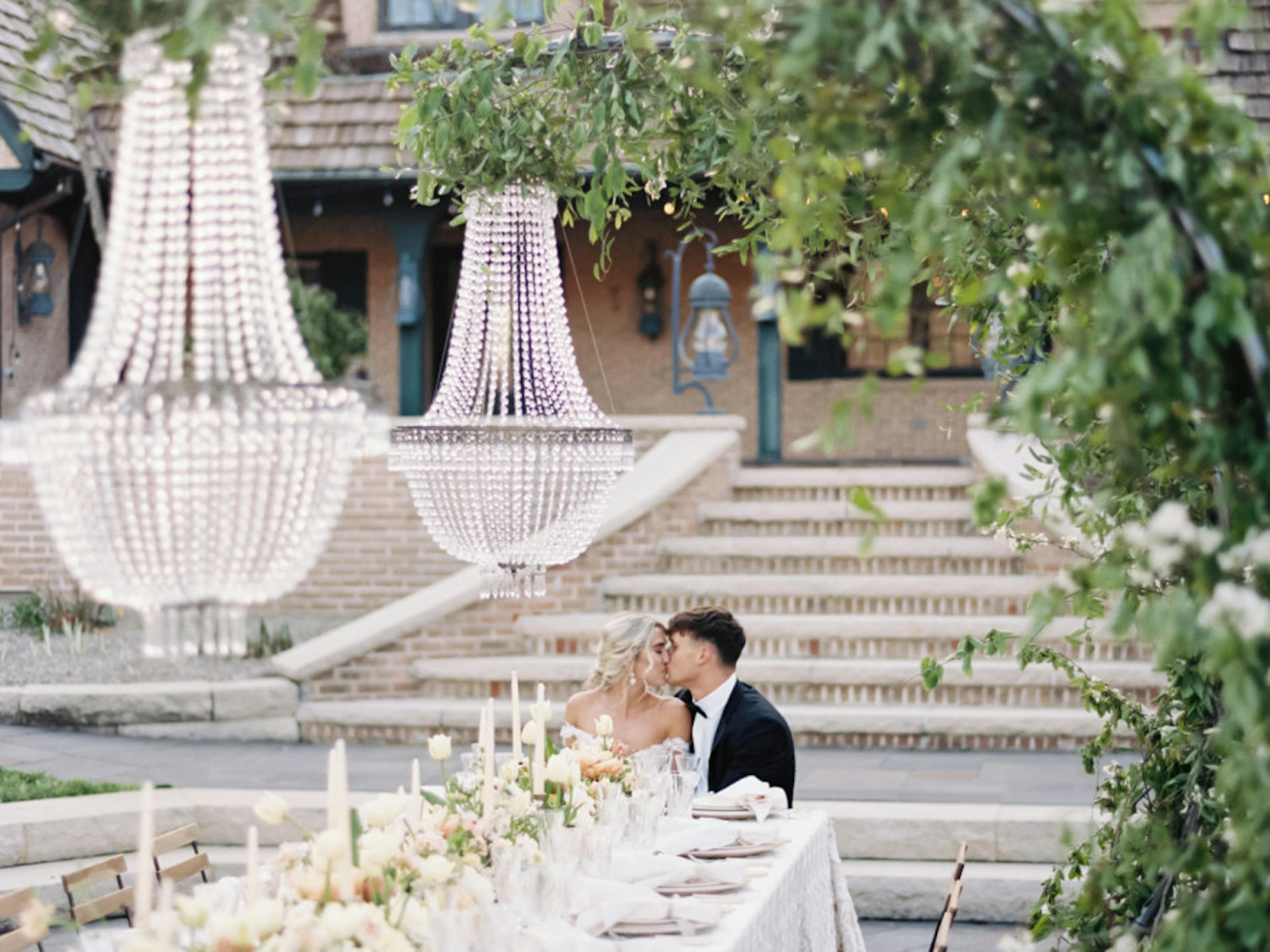 Bride and groom kiss at the end of a banquet table in that garden at modern European themed outdoor wedding with neutral color palette.