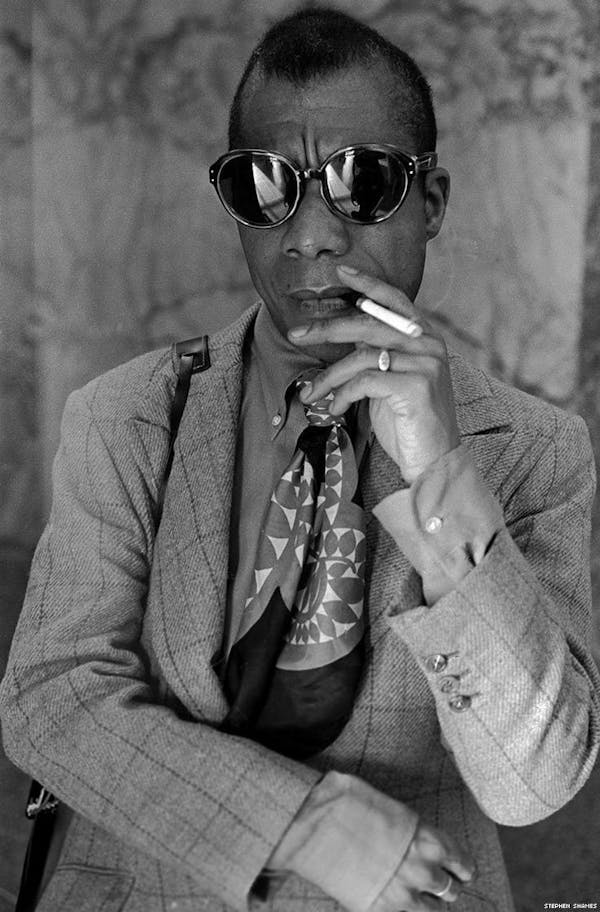 James Baldwin fashion style in a suit, neck scarf, and sunglasses smoking a cigarette.