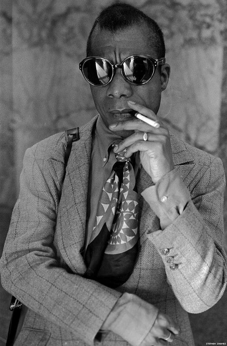 James Baldwin fashion style in a suit, neck scarf, and sunglasses smoking a cigarette.