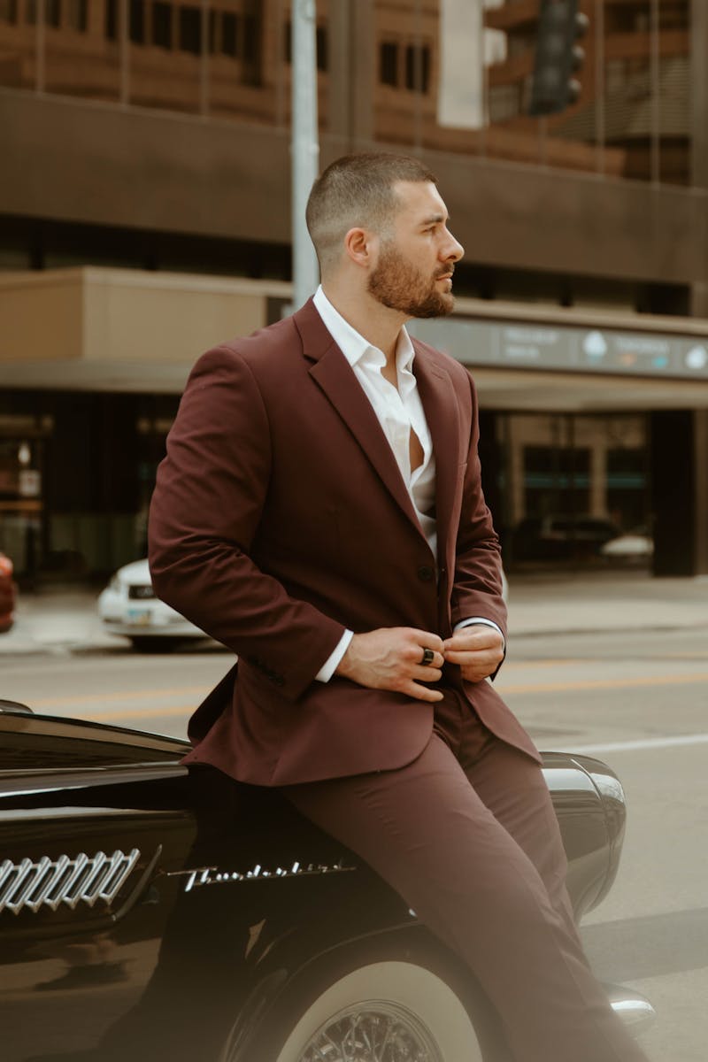 Man wearing red suit with no tie and white dress shirt leaning on hood of a sports car.