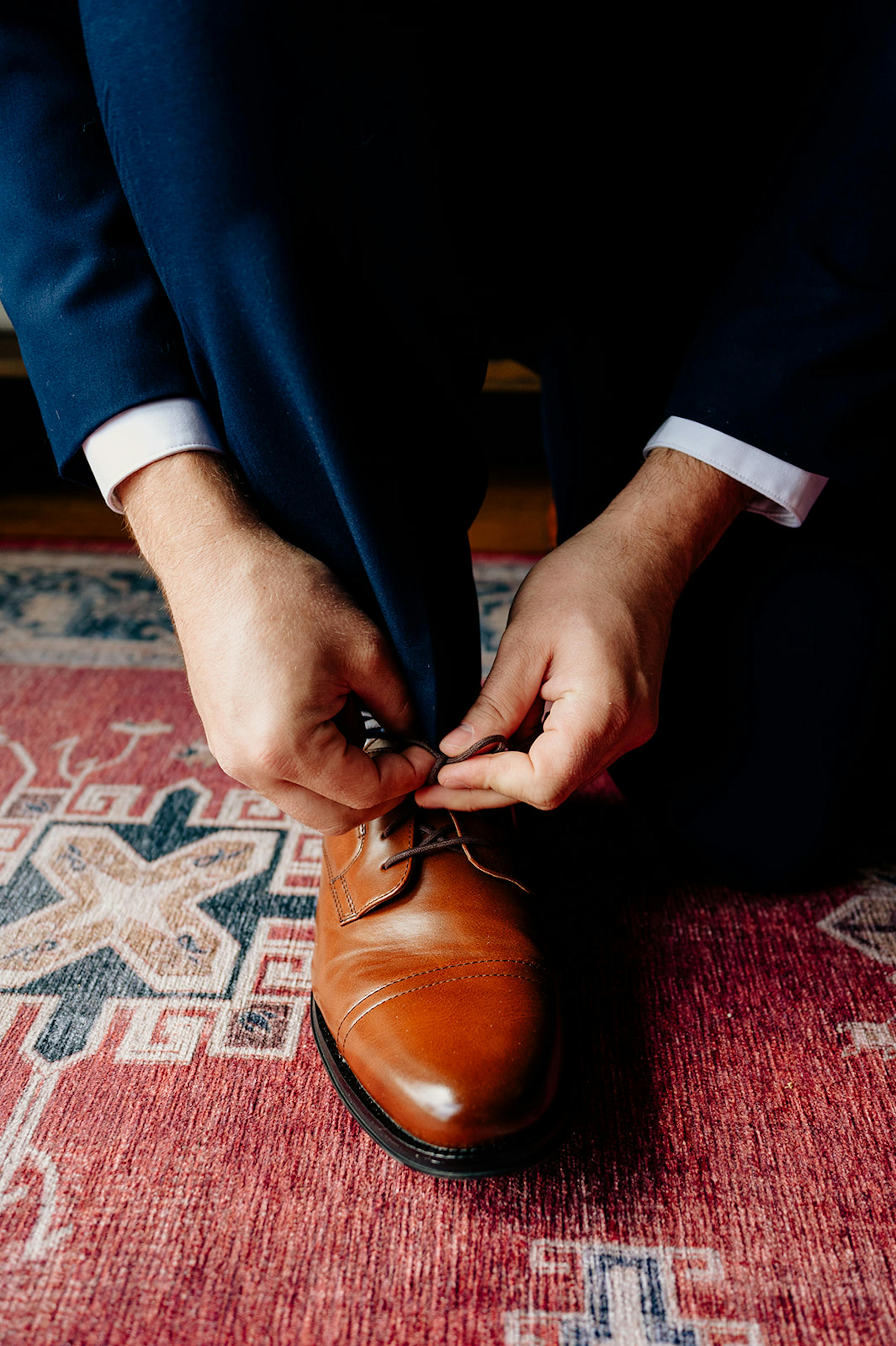 Man wearing navy blue suit tying cognac brown dress shoe on venue rug