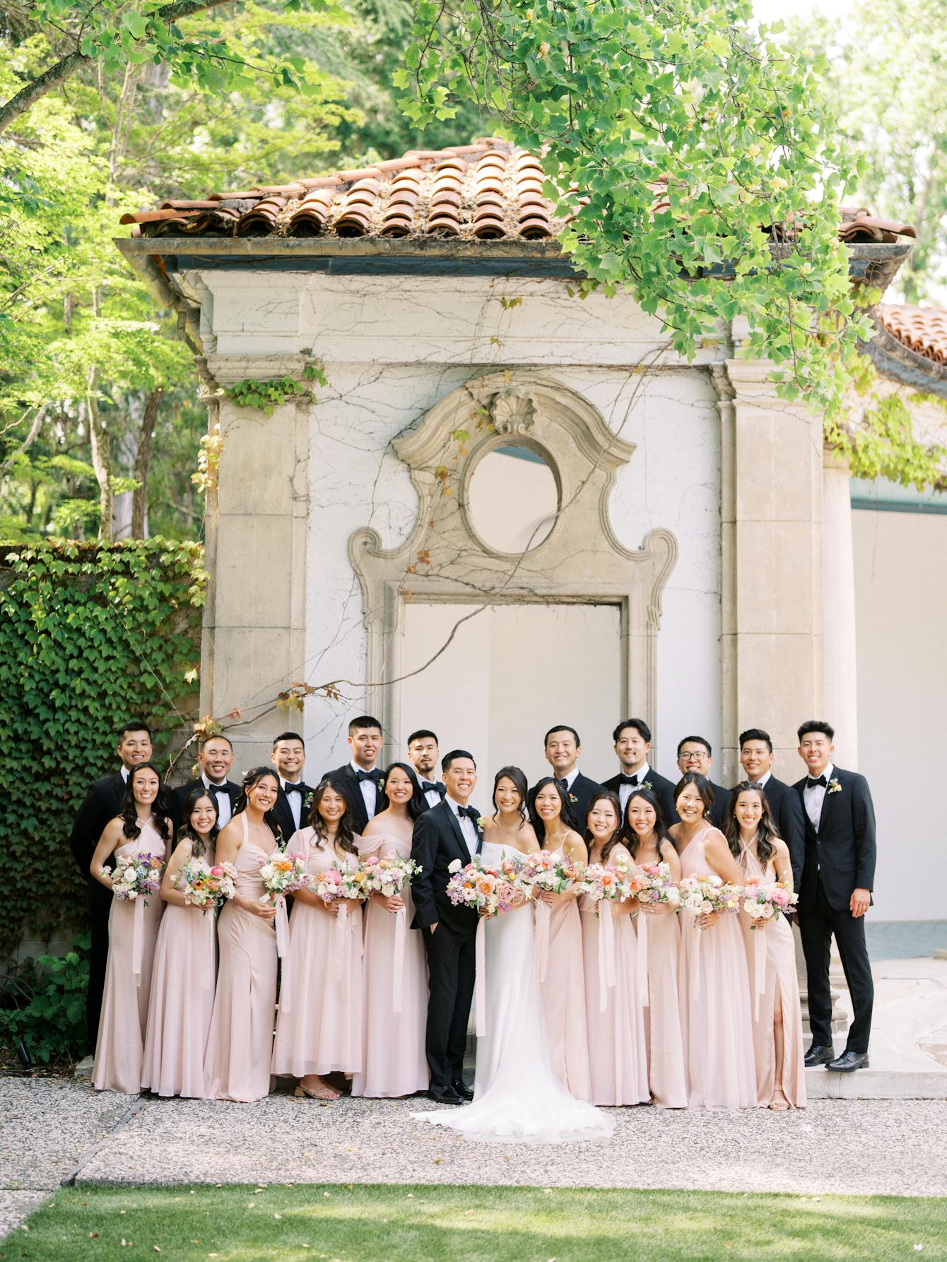Wedding couple surrounded by groomsmen in black suits and bridesmaids in plum pink.