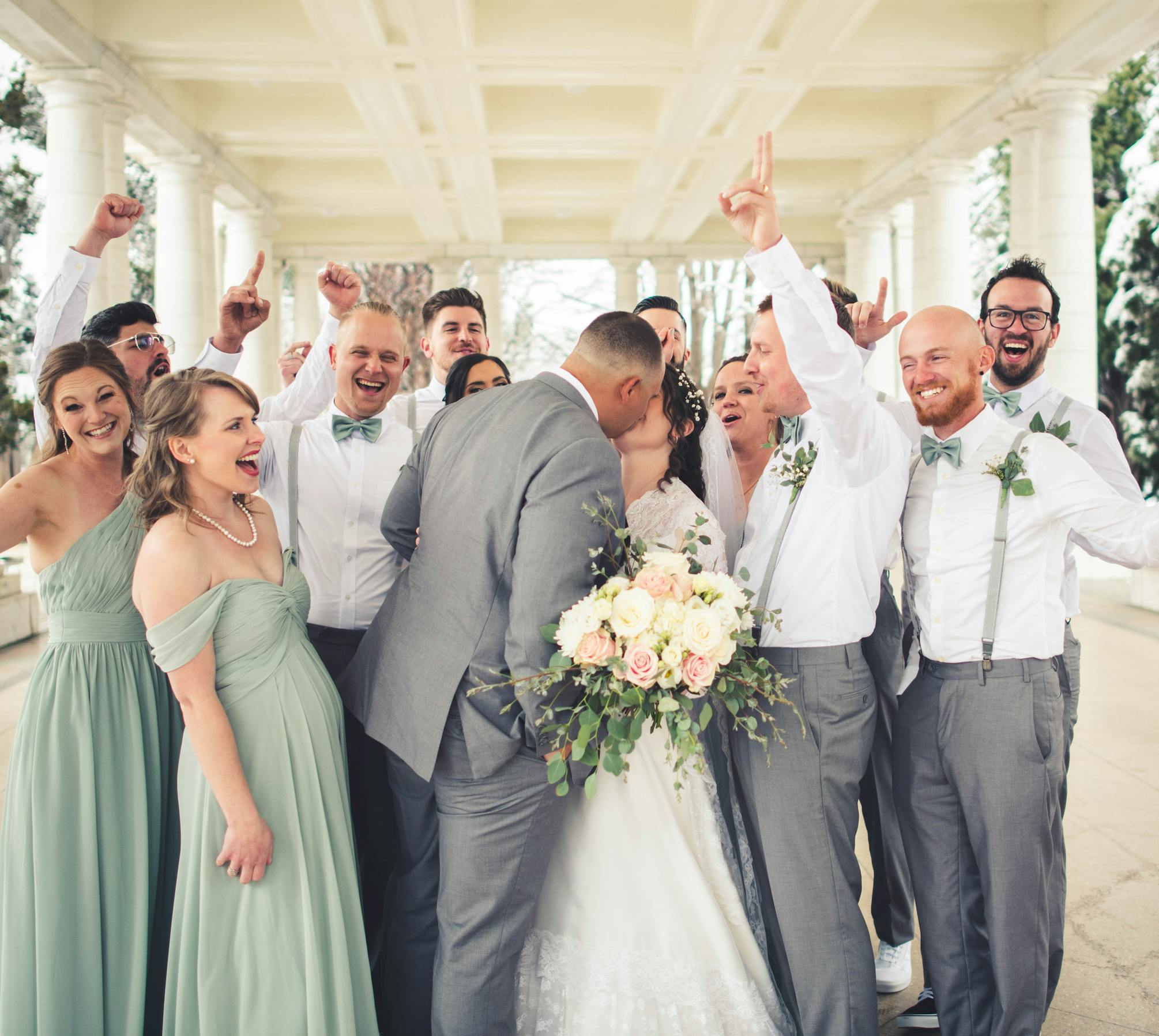 Kissing wedding couple celebrated by bridesmaids in olive green dresses and groomsmen in white shirts and textured gray suit pants.