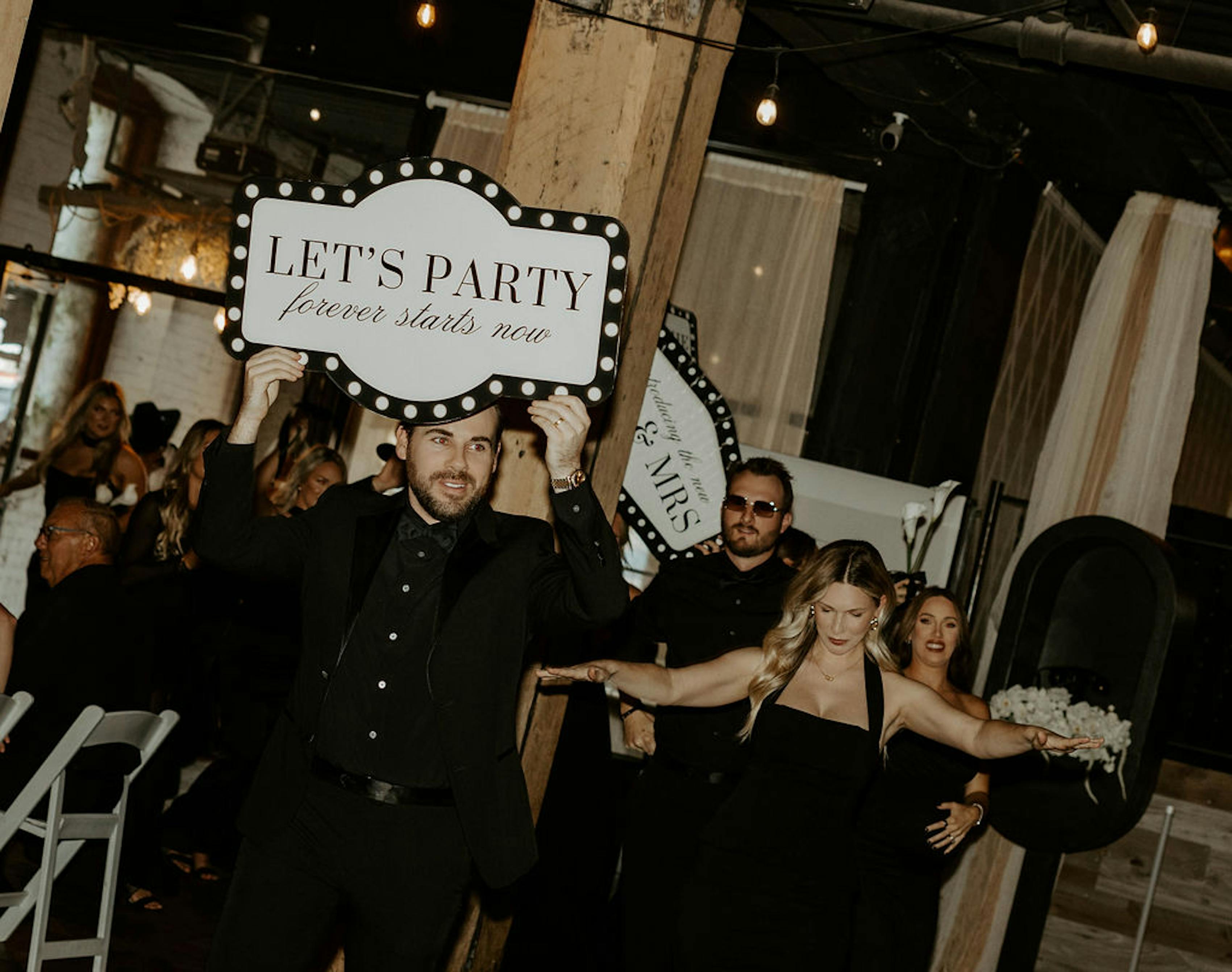 groomsman in all-black outfit holding fun wedding party entrance sign
