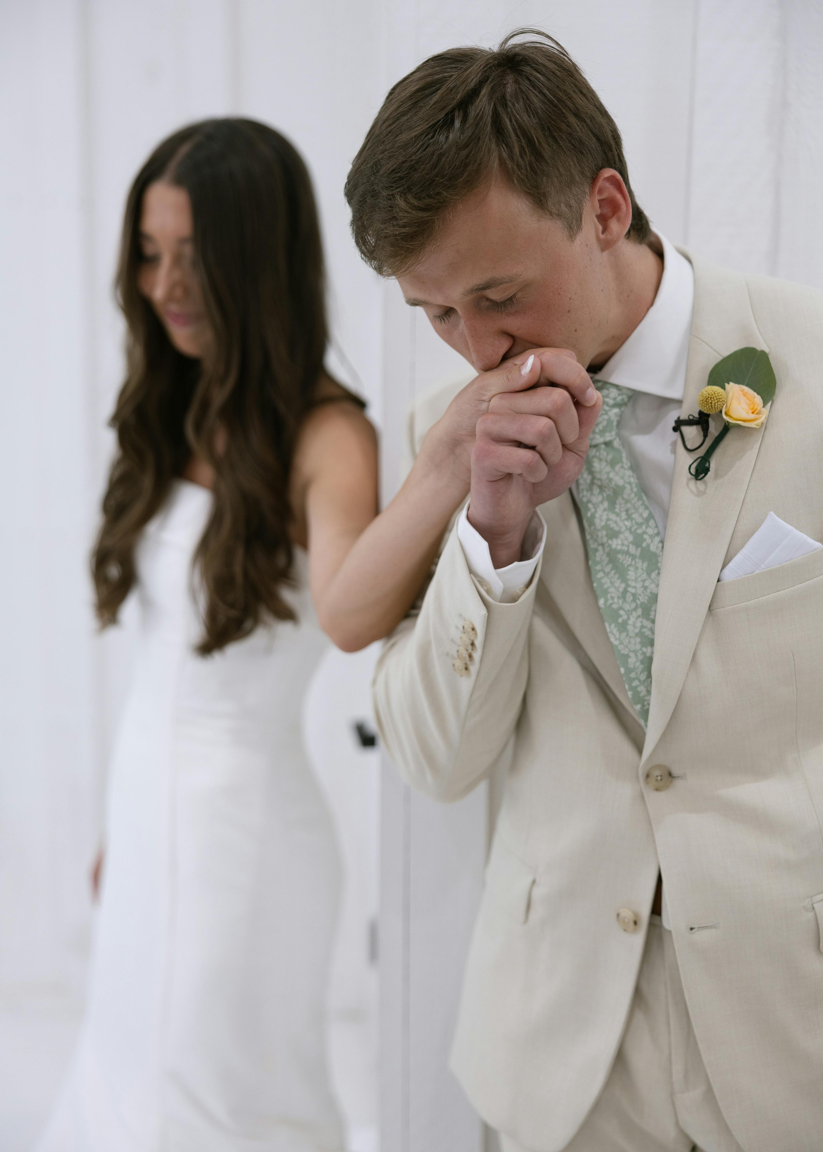 Groom wearing tan suit kissing bride's hand at rustic wedding
