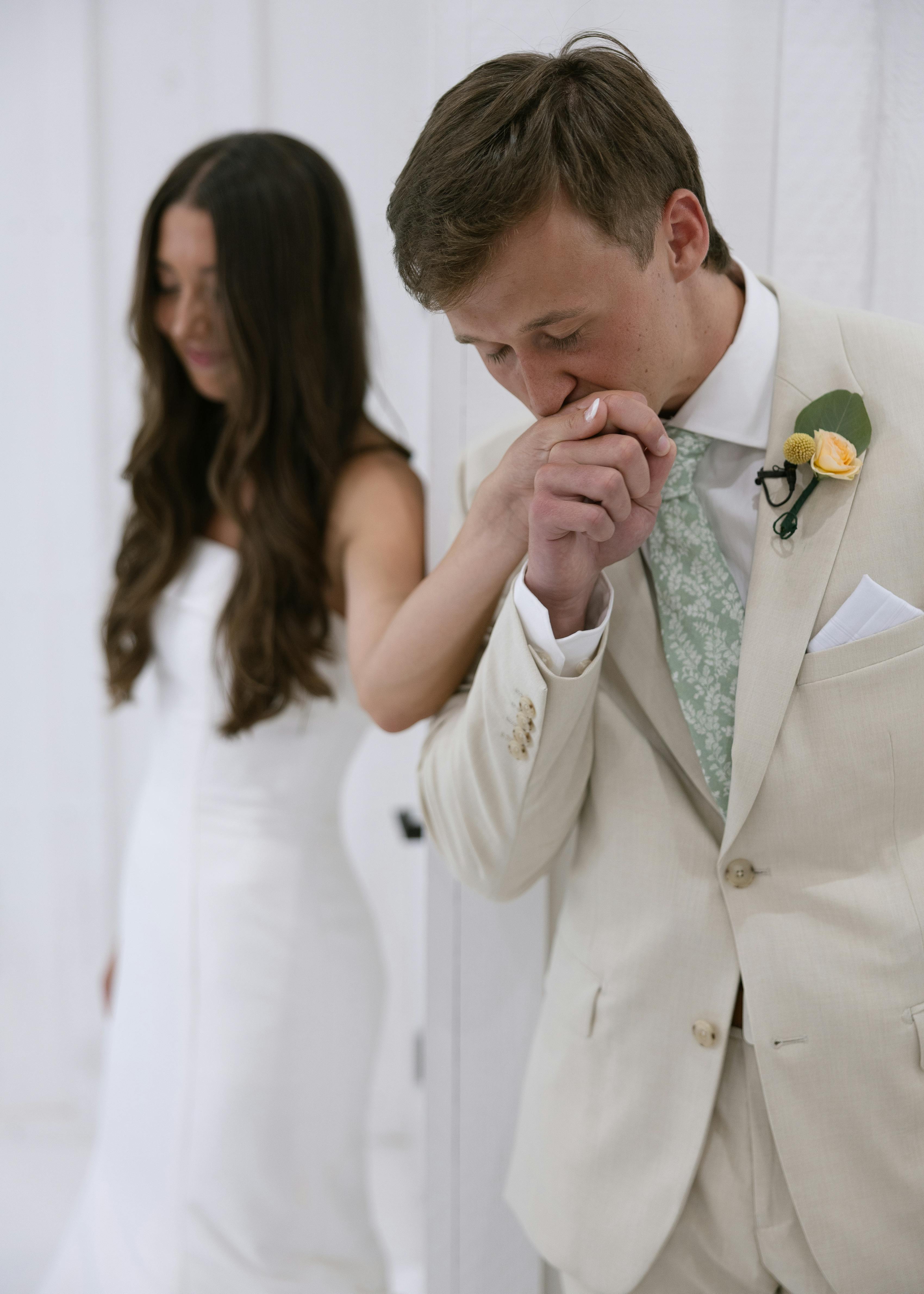 Groom wearing tan suit kissing bride's hand at rustic wedding