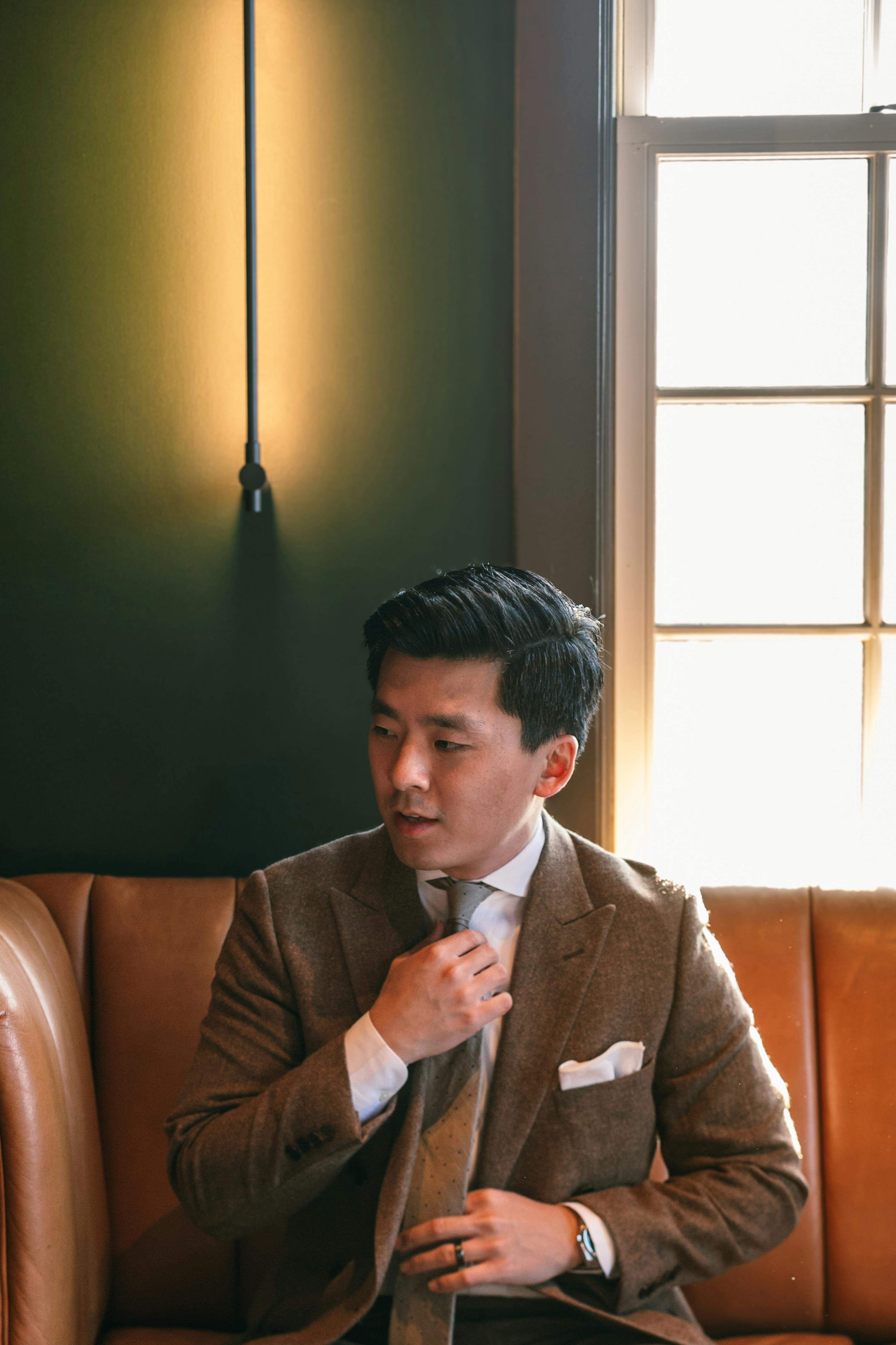 Man wearing brown tweed blazer sitting on couch adjusting tie