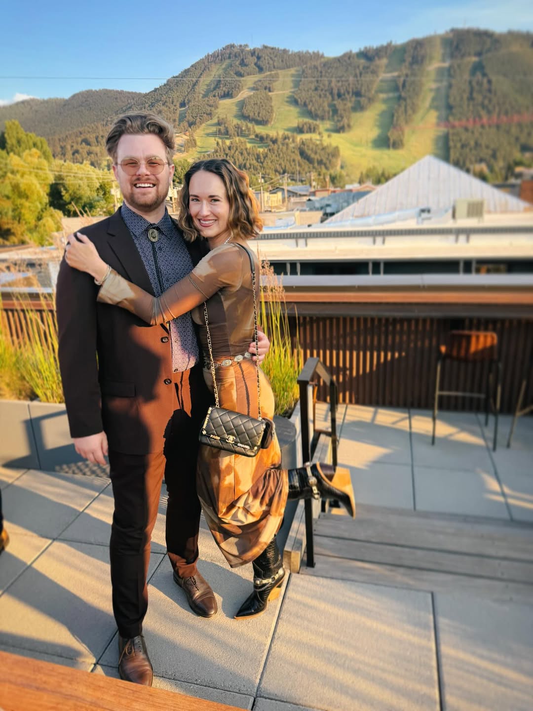 Couple attending mountain rehearsal dinner wearing dark brown suit and bolo tie and brown dress