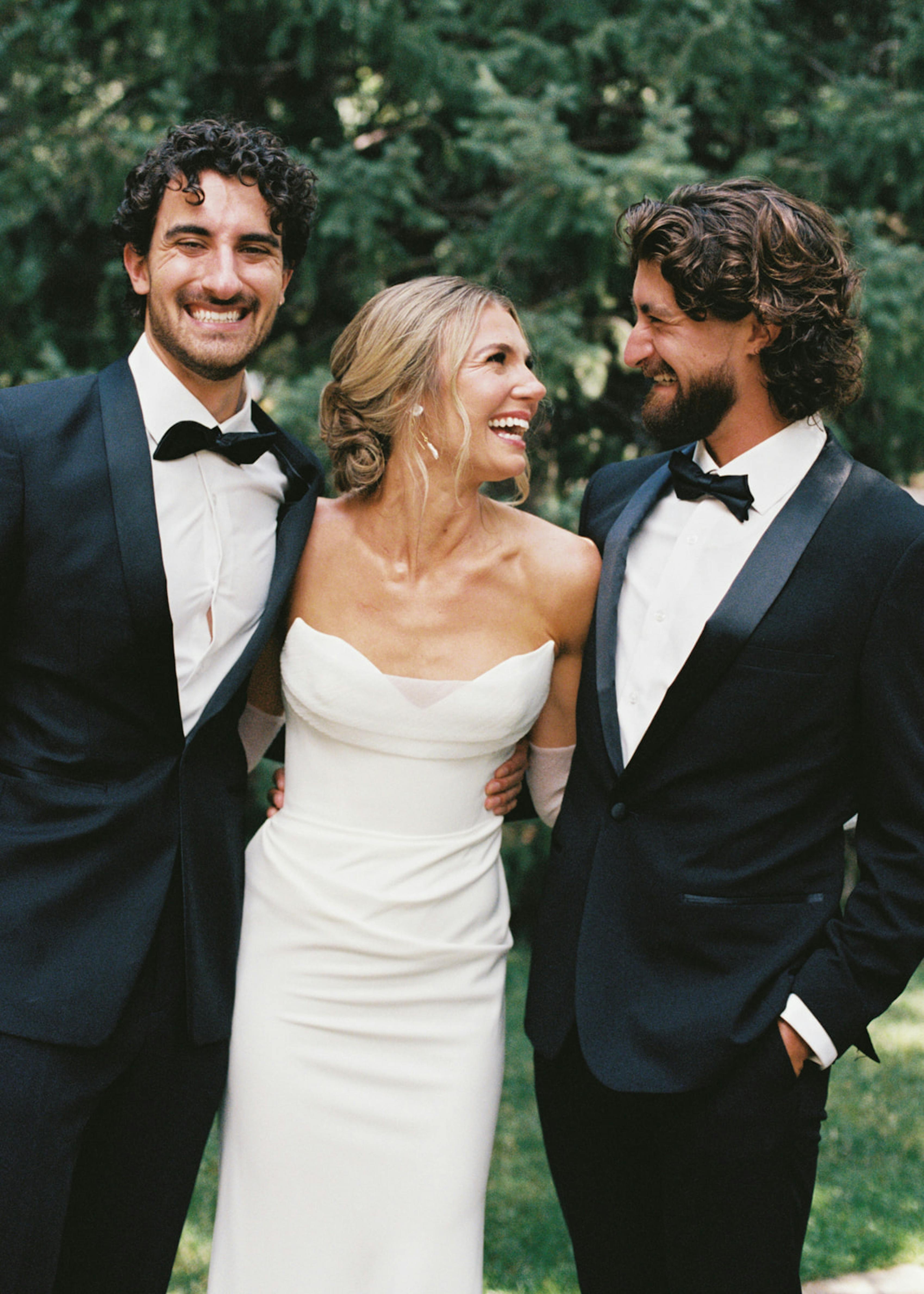 Bride wearing strapless wedding gown hugging brothers wearing black tuxedos