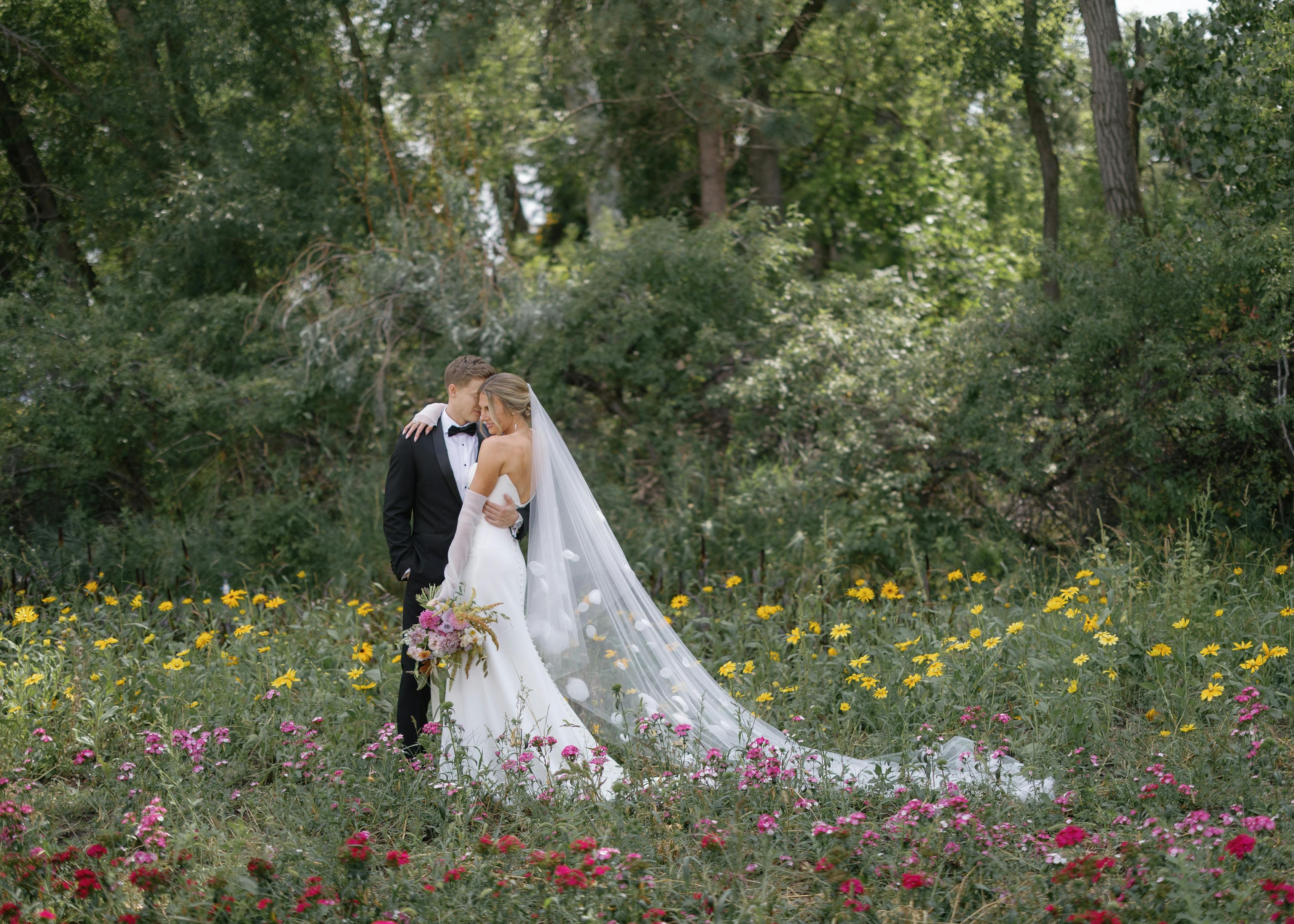 Intimate bride and groom portrait with black tuxedo, gown, and floral veil in wildflower field