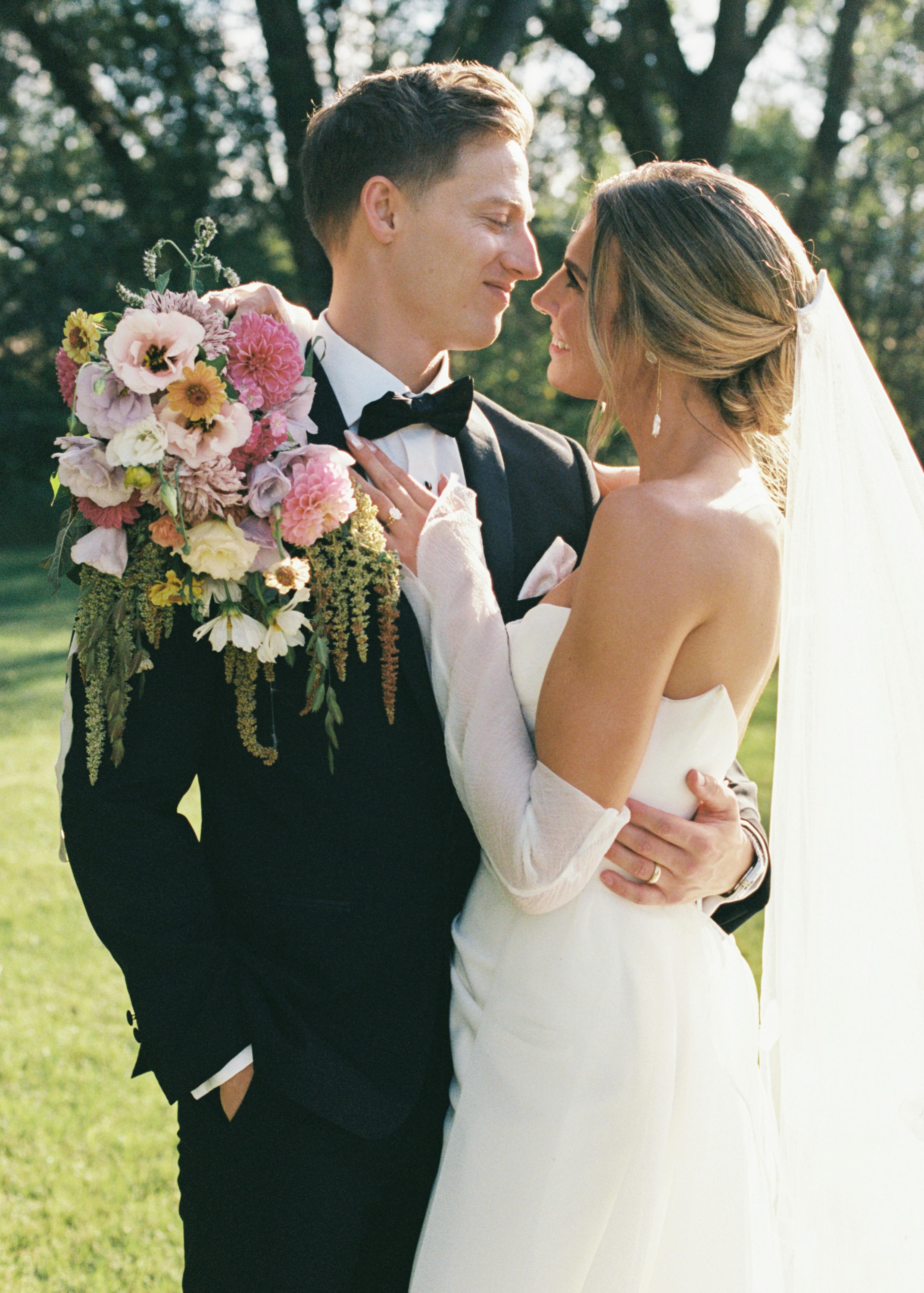 Bride wearing sheer gloves hugging groom in black tuxedo posing with modern pastel bouquet