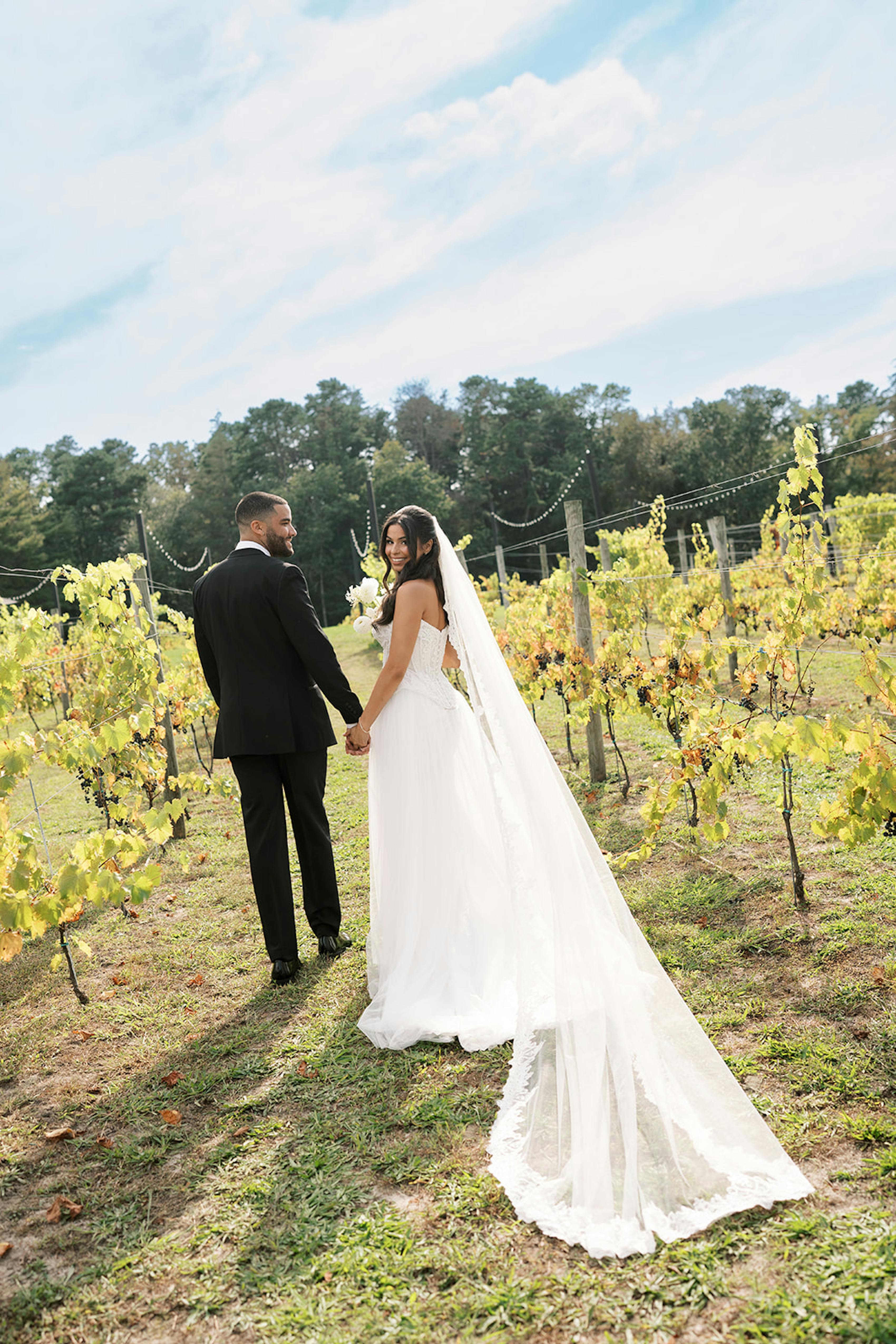 Bride wearing tulle dress and groom in tuxedo walking through vineyard
