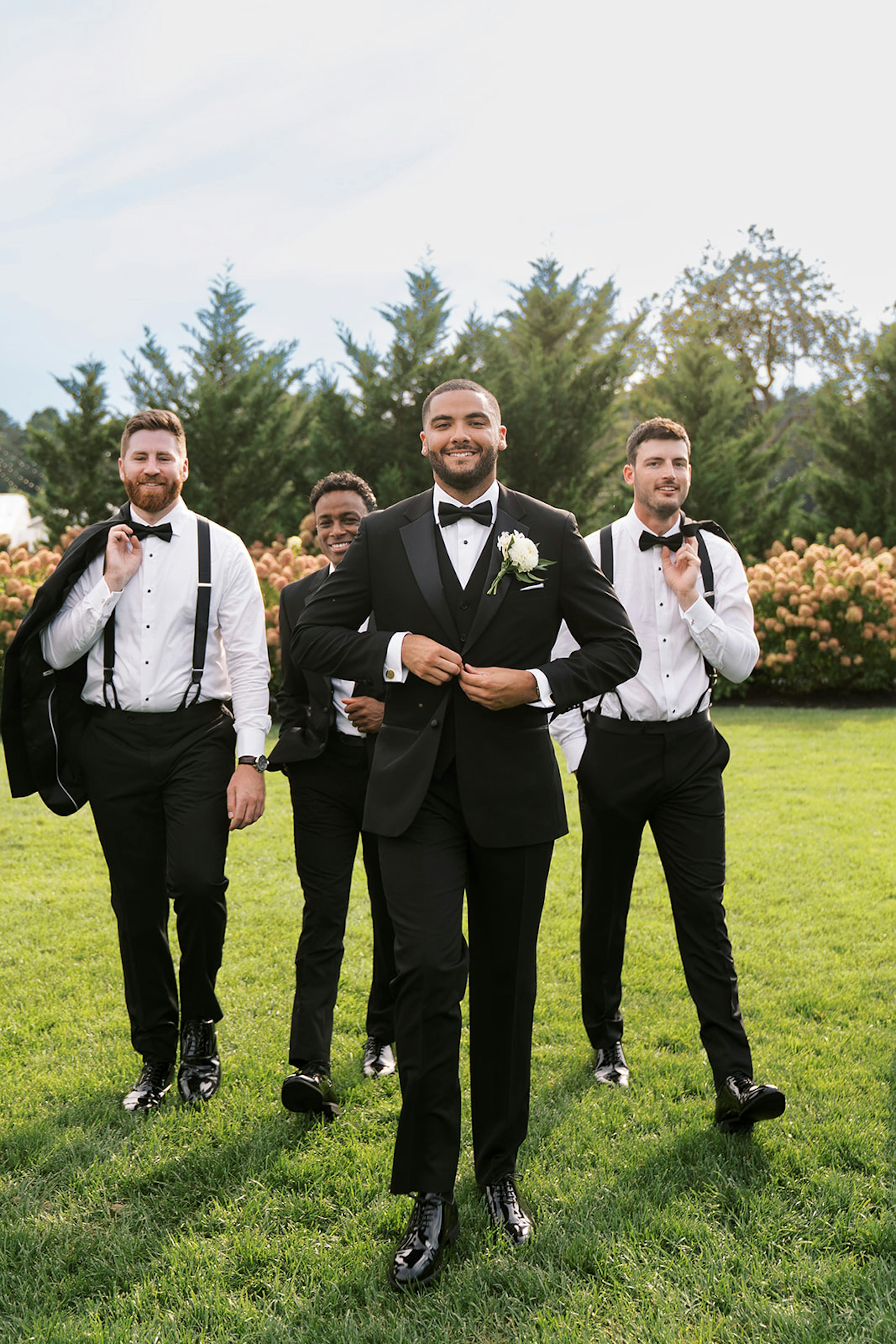 Groom wearing 3 piece black tuxedo walking with groomsmen wearing tux pants and suspenders