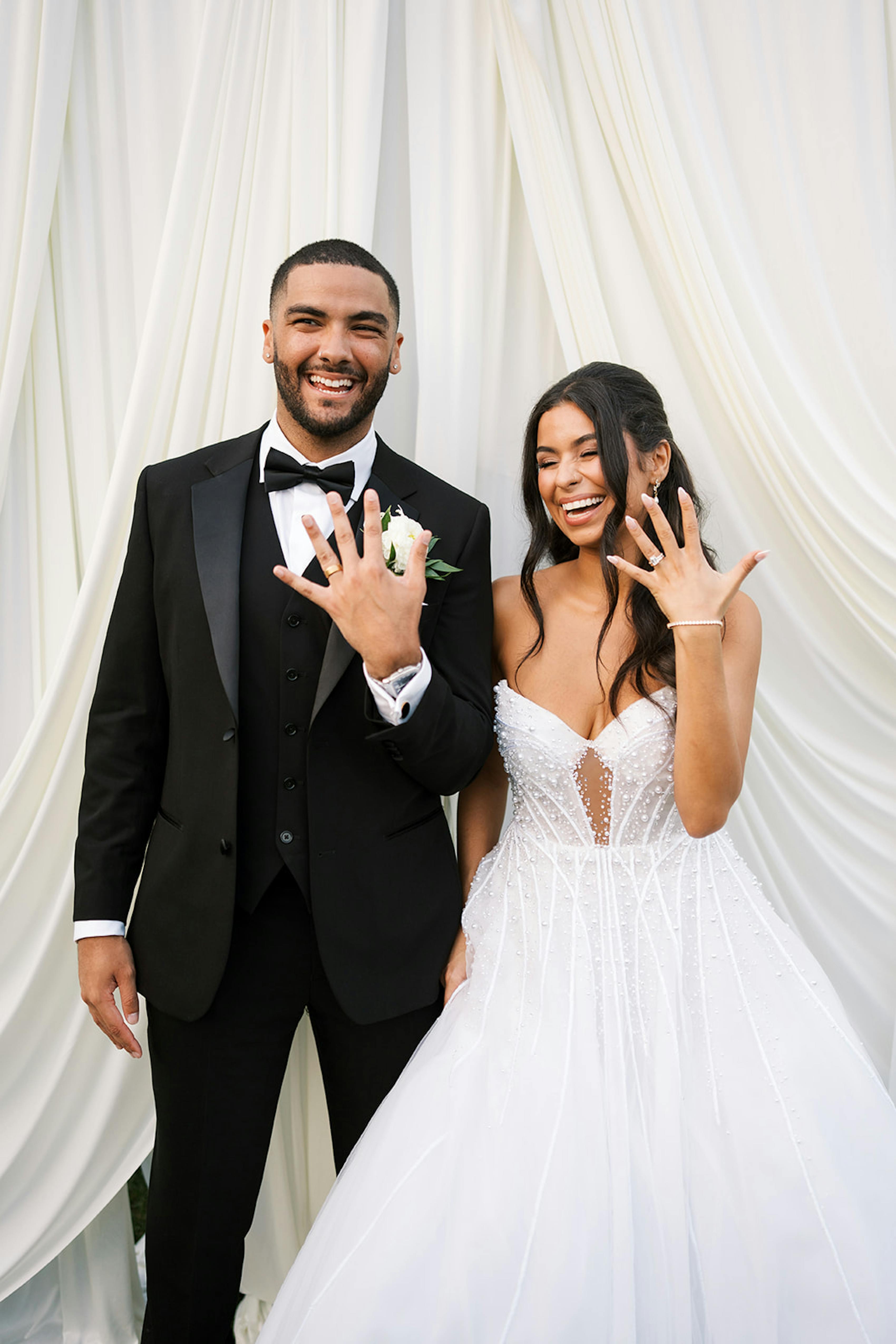 Bride and groom at modern black tie wedding showing off rings in front of draped fabric backdrop