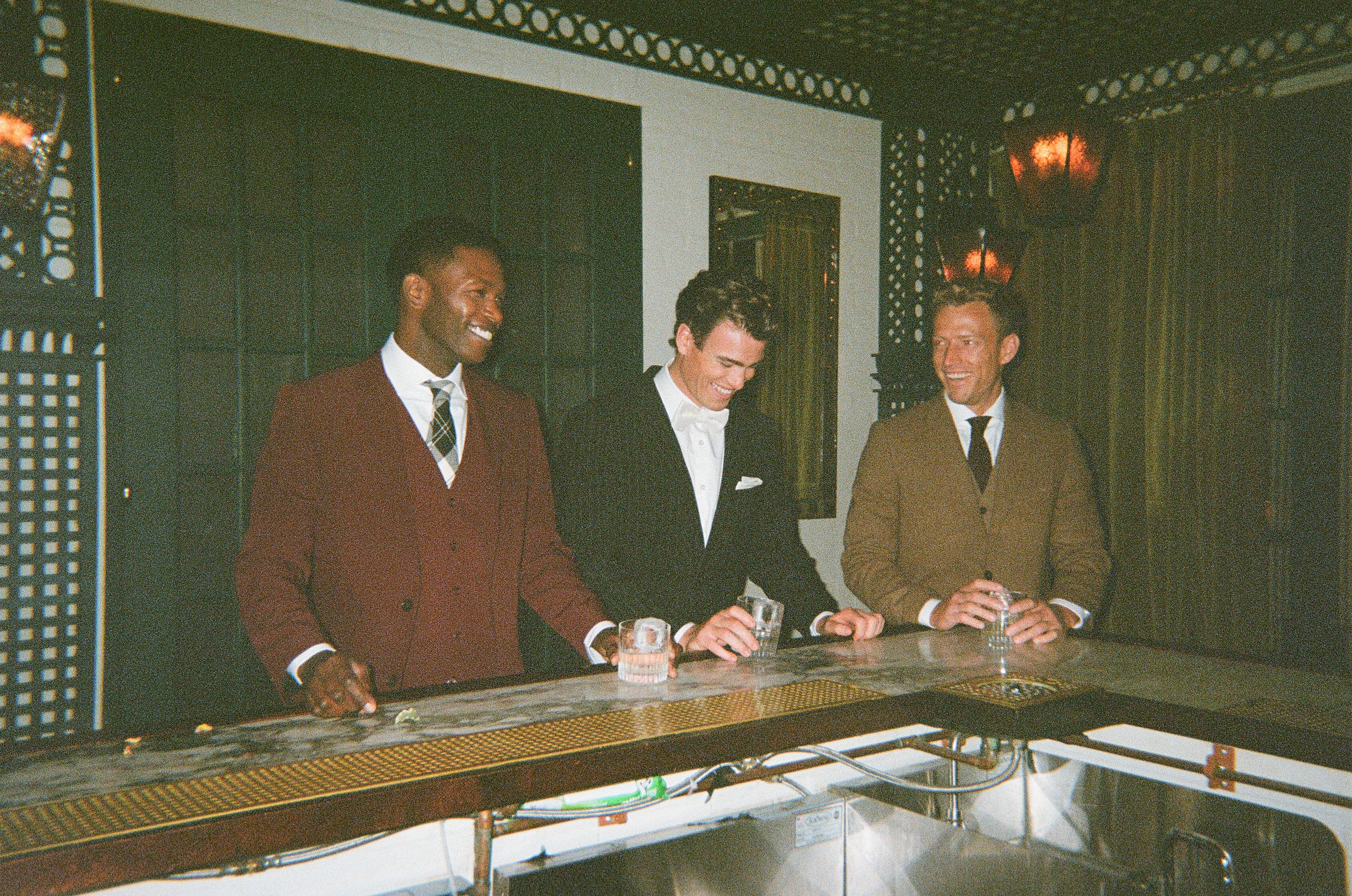 3 guys at bar wearing wedding guest outfits with burgundy suit, black suit, and tweed suit