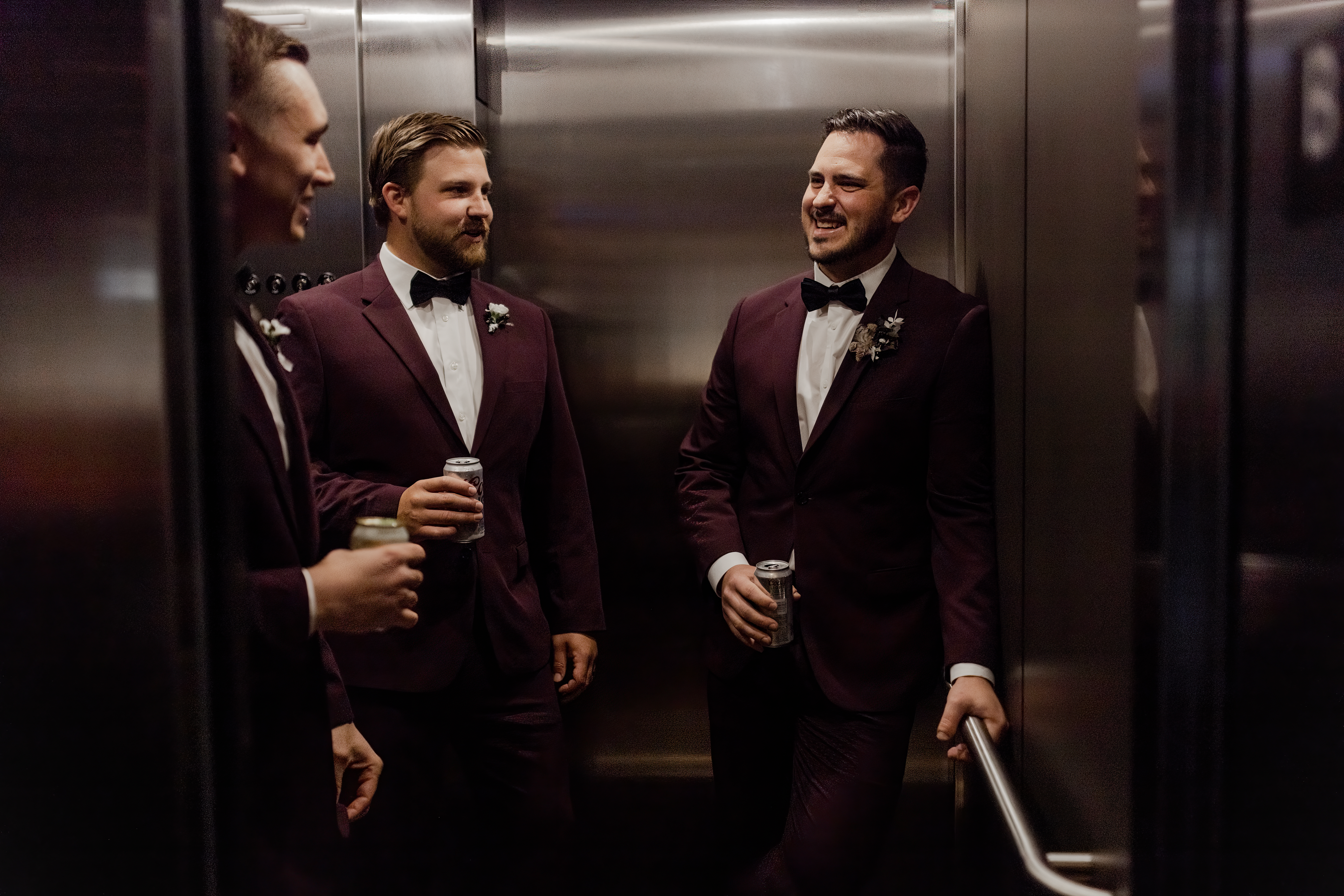 Groomsmen holding beers in elevator wearing burgundy wedding suits and bow ties