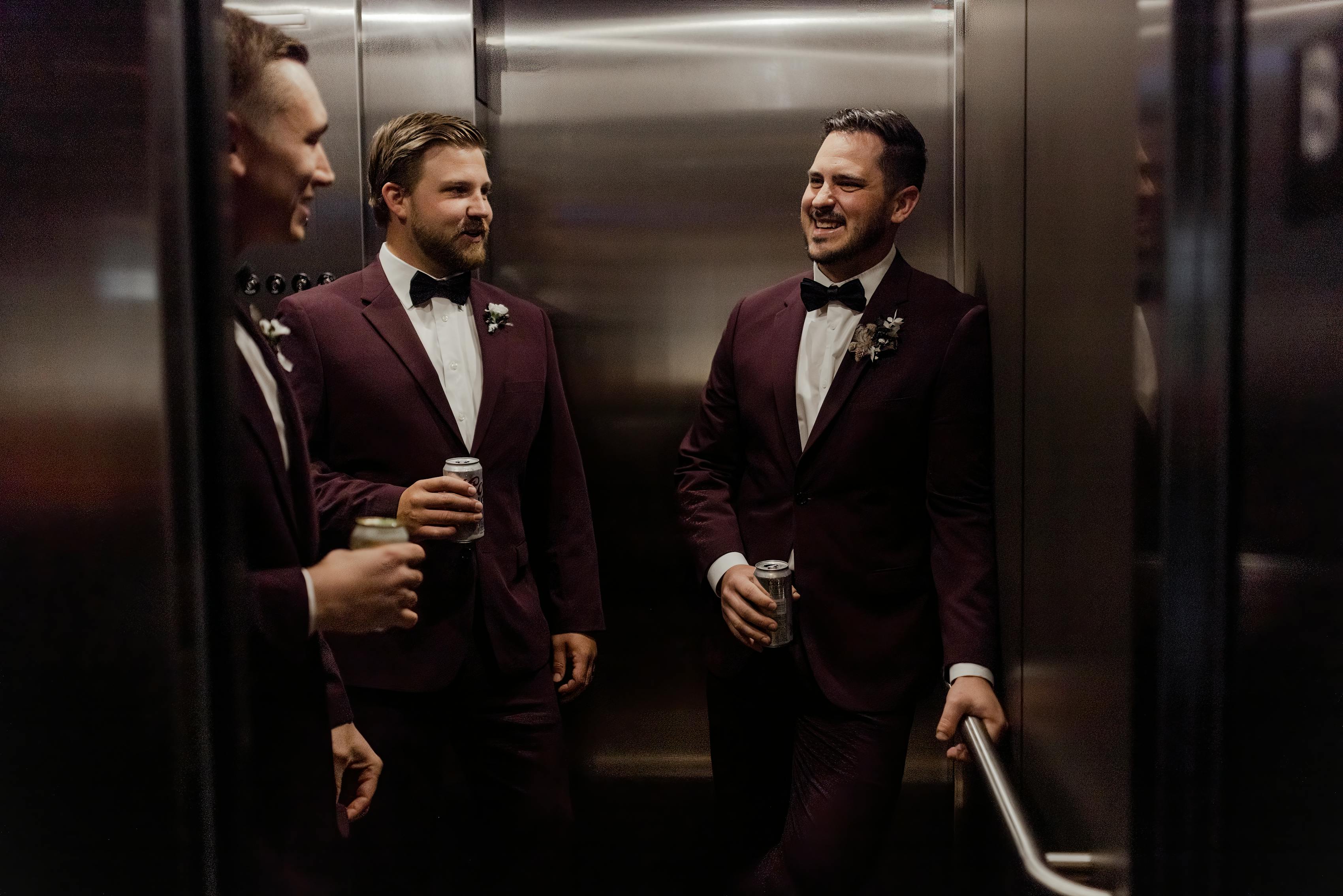 Groomsmen holding beers in elevator wearing burgundy wedding suits and bow ties