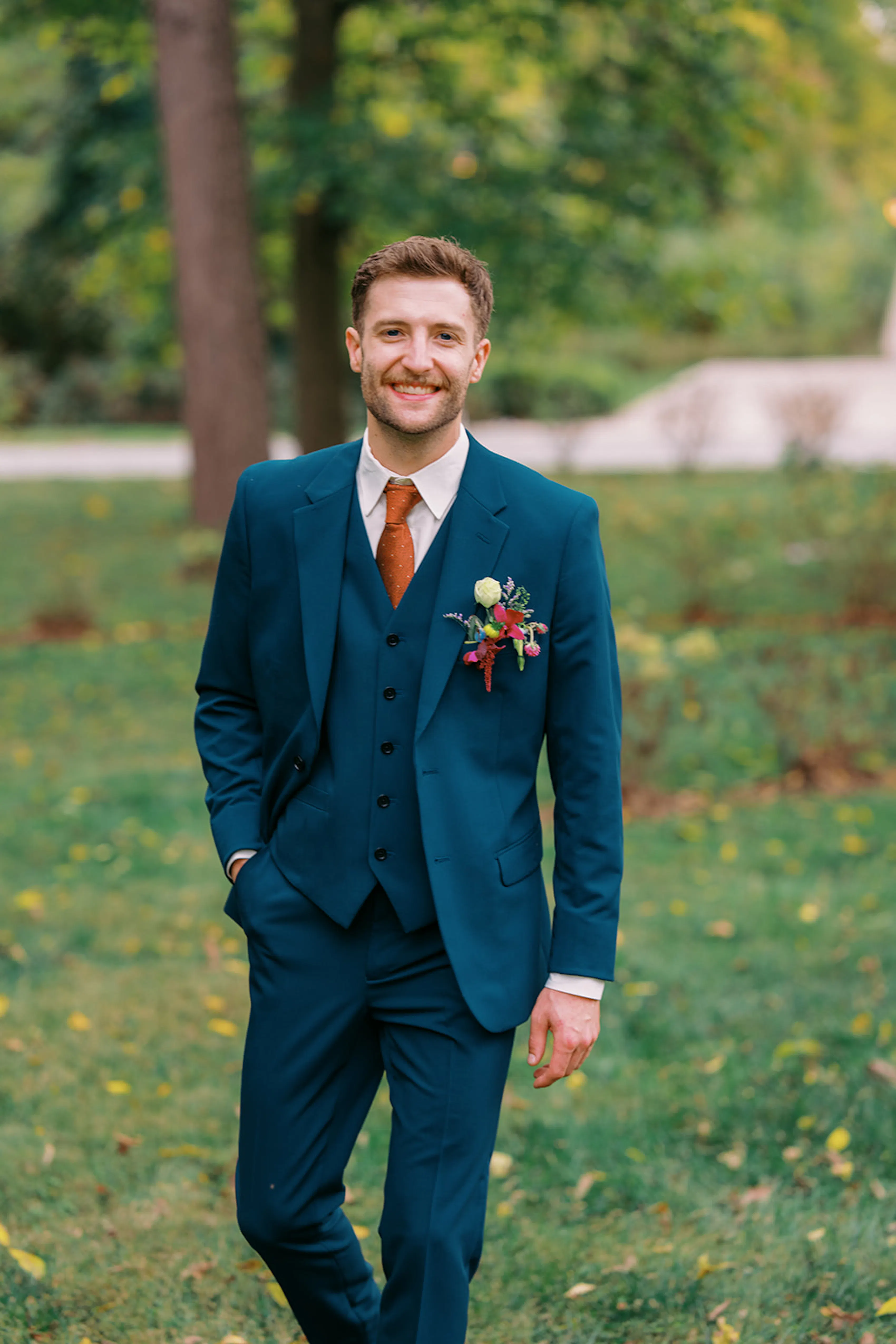 Groom outfit with 3-piece deep teal suit, colorful boutonniere, and rust orange tie